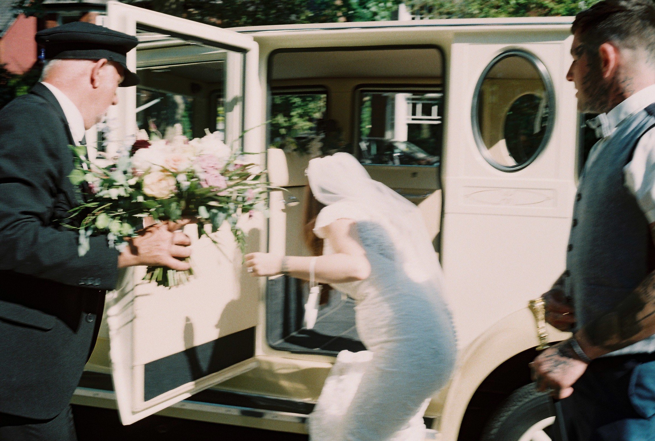 Bride in a white dress and veil enters a vintage car with two men, one holding a bouquet of flowers, outside a building with trees in the background.