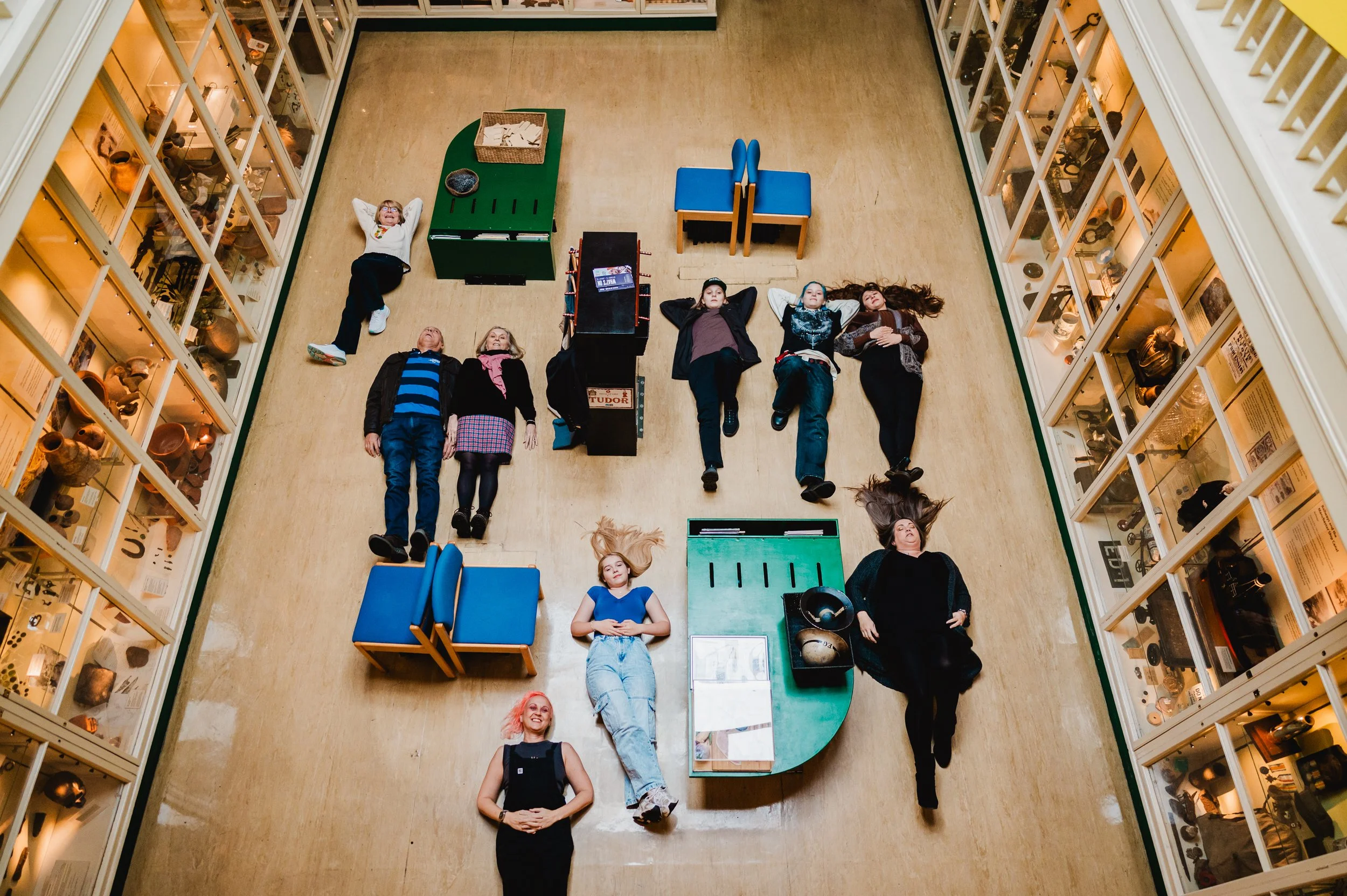 People lying on the floor of a museum or gallery, viewed from above, surrounded by display cases with artifacts.