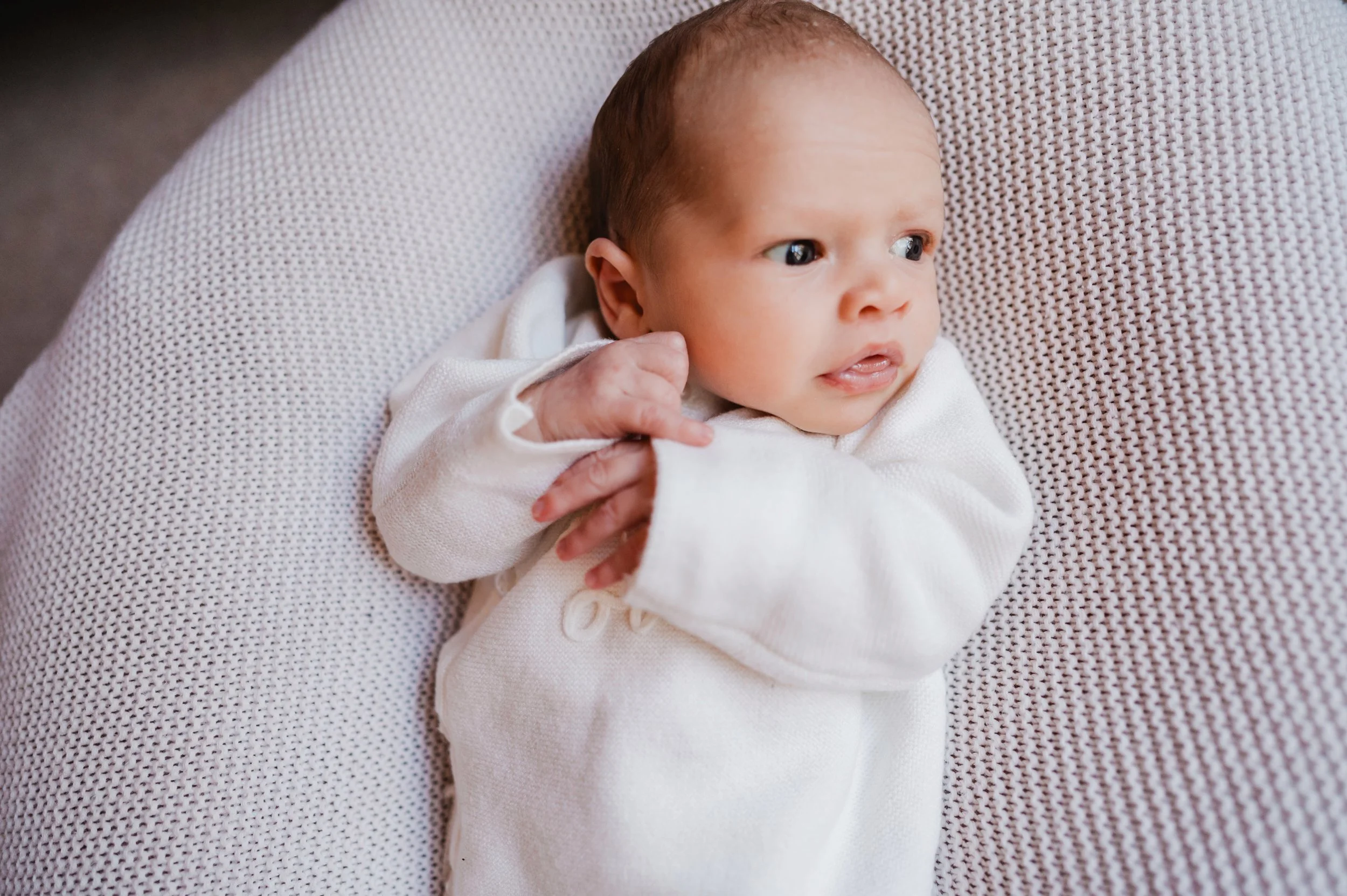 Close-up of a baby lying on a textured, light-colored surface, wearing a white outfit and looking to the side with an attentive expression.