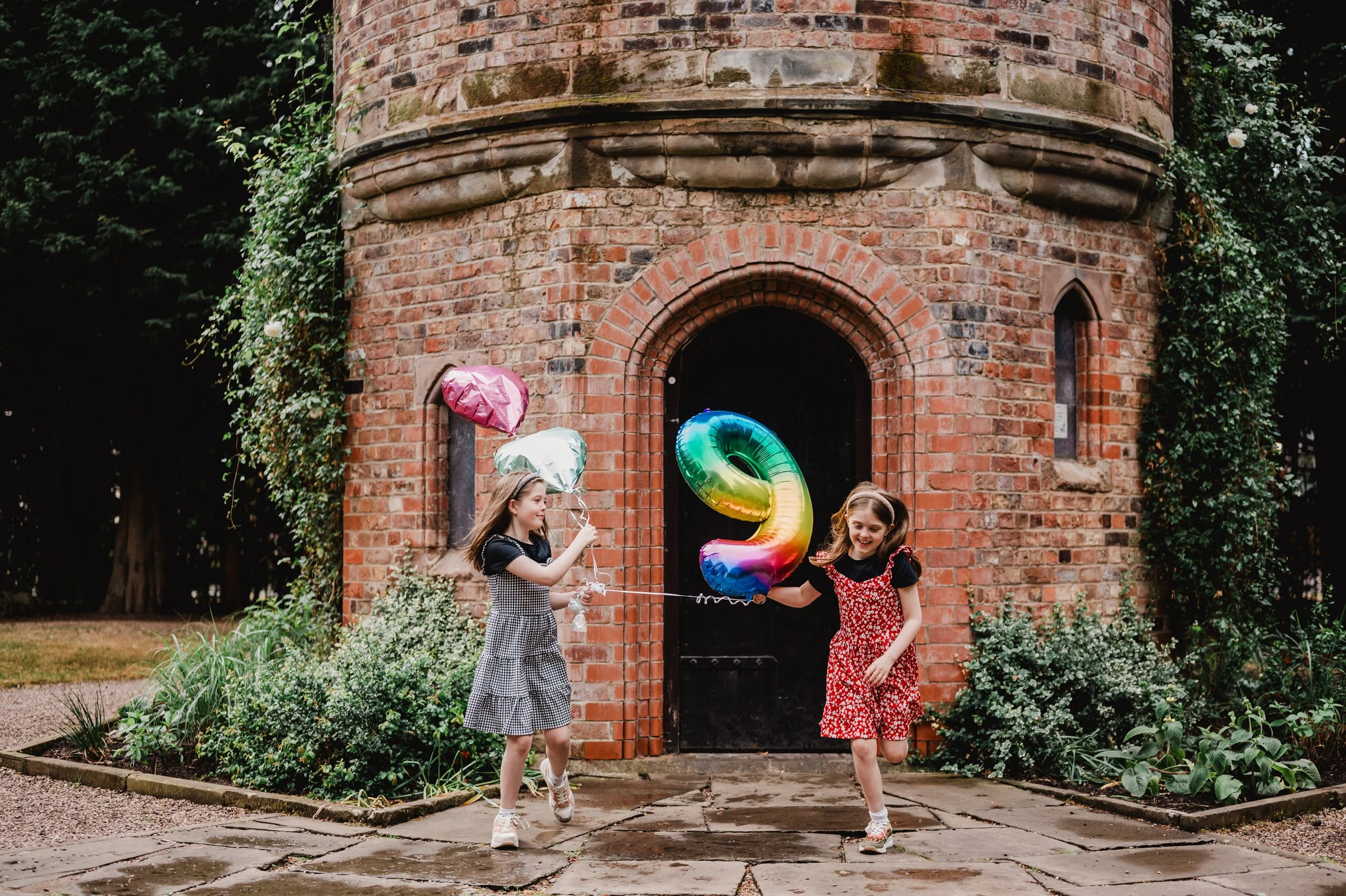 Two young girls holding colorful balloons walking in front of a brick building with a black door and surrounded by greenery.