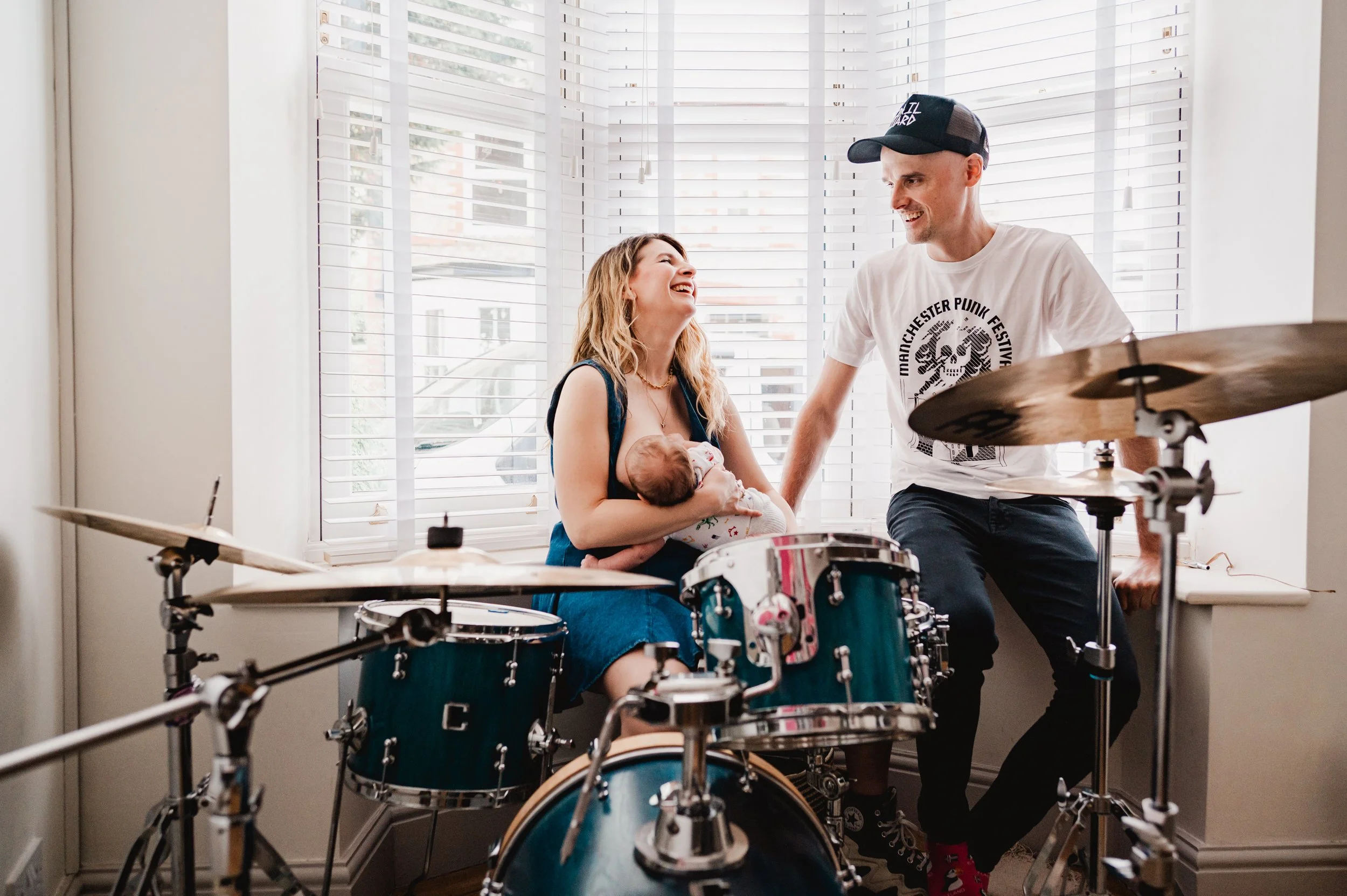 A woman holding a baby giggles as a man smiles at her, standing next to a drum set in a bright room with large windows and white blinds.