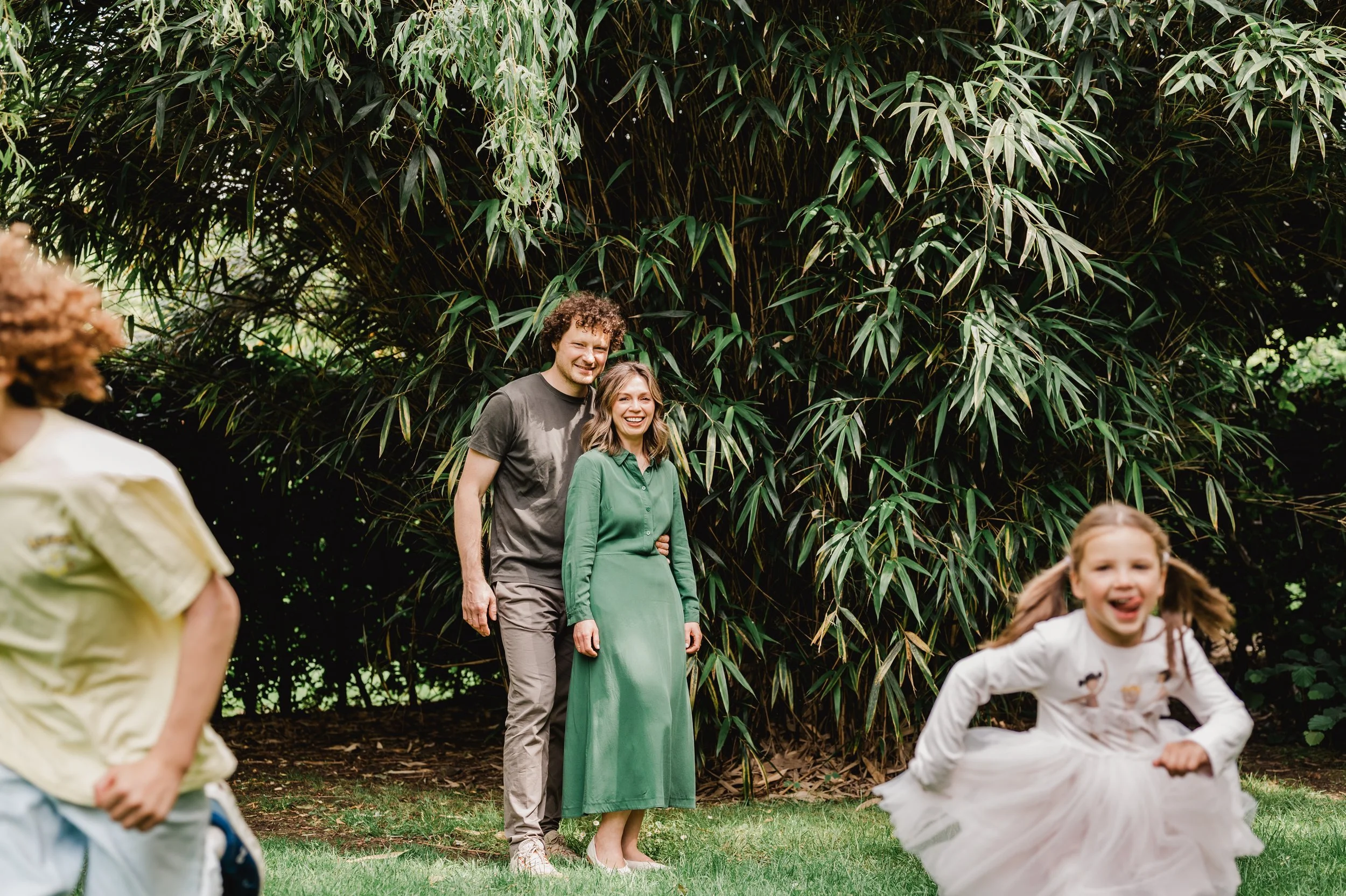 A group of children and a woman outdoors in front of tall green bamboo plants, with children running in a playful manner.