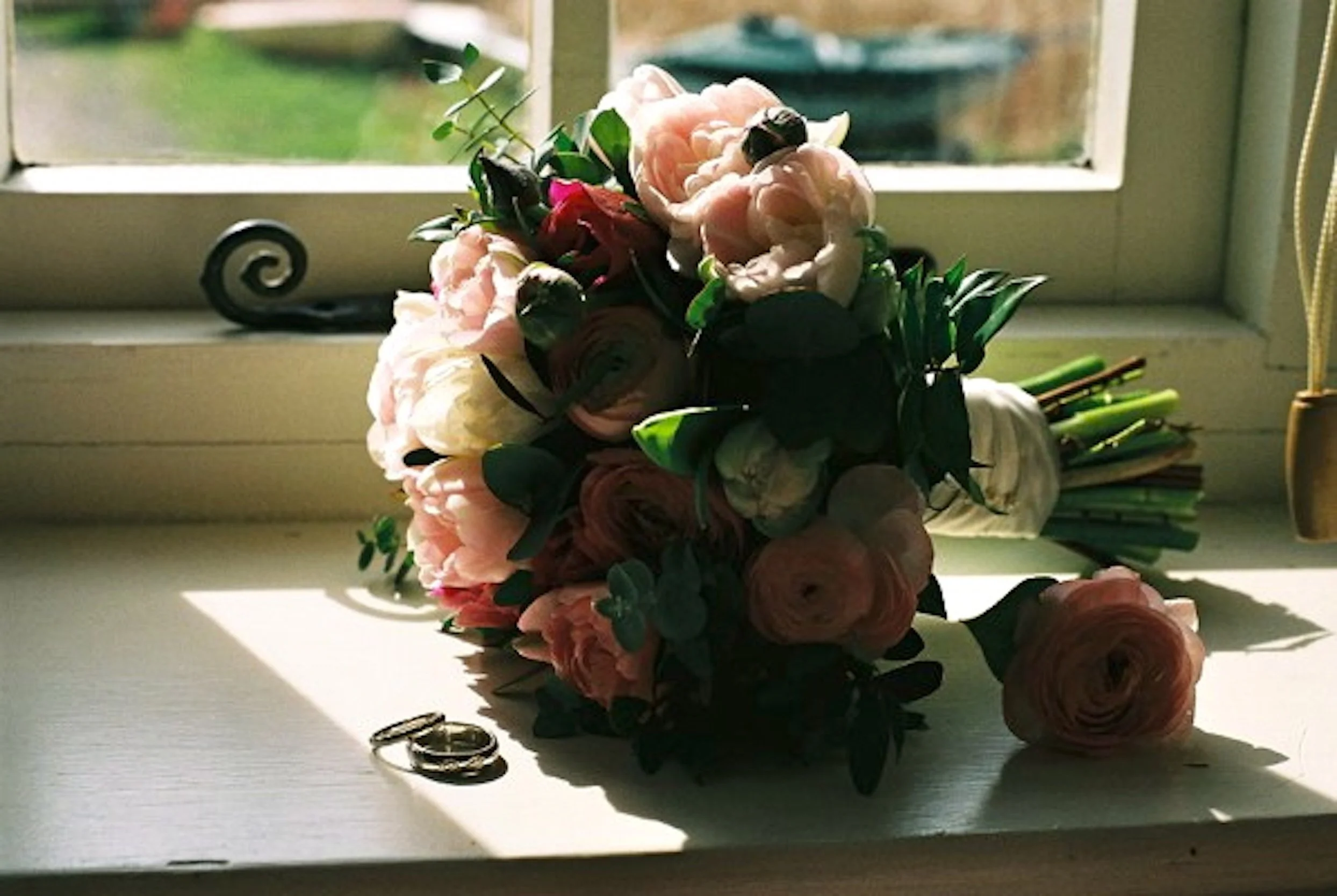 A bouquet of pink, white, and peach flowers with green leaves and stems, resting on a windowsill with sunlight streaming through the window, casting shadows.
