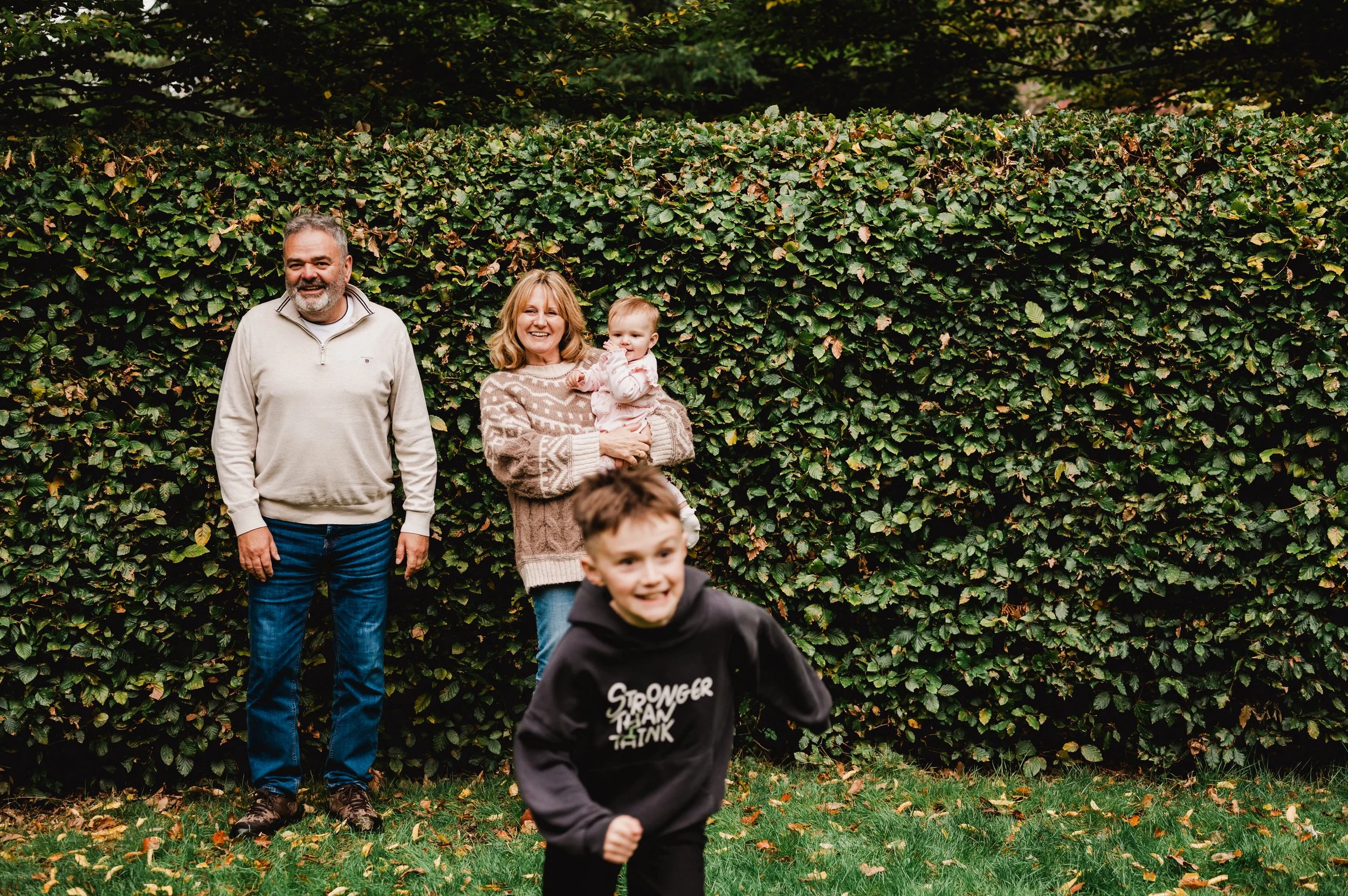 A group of children and two adults standing and playing outdoors near a large hedge of green leaves with some fallen leaves on the ground.