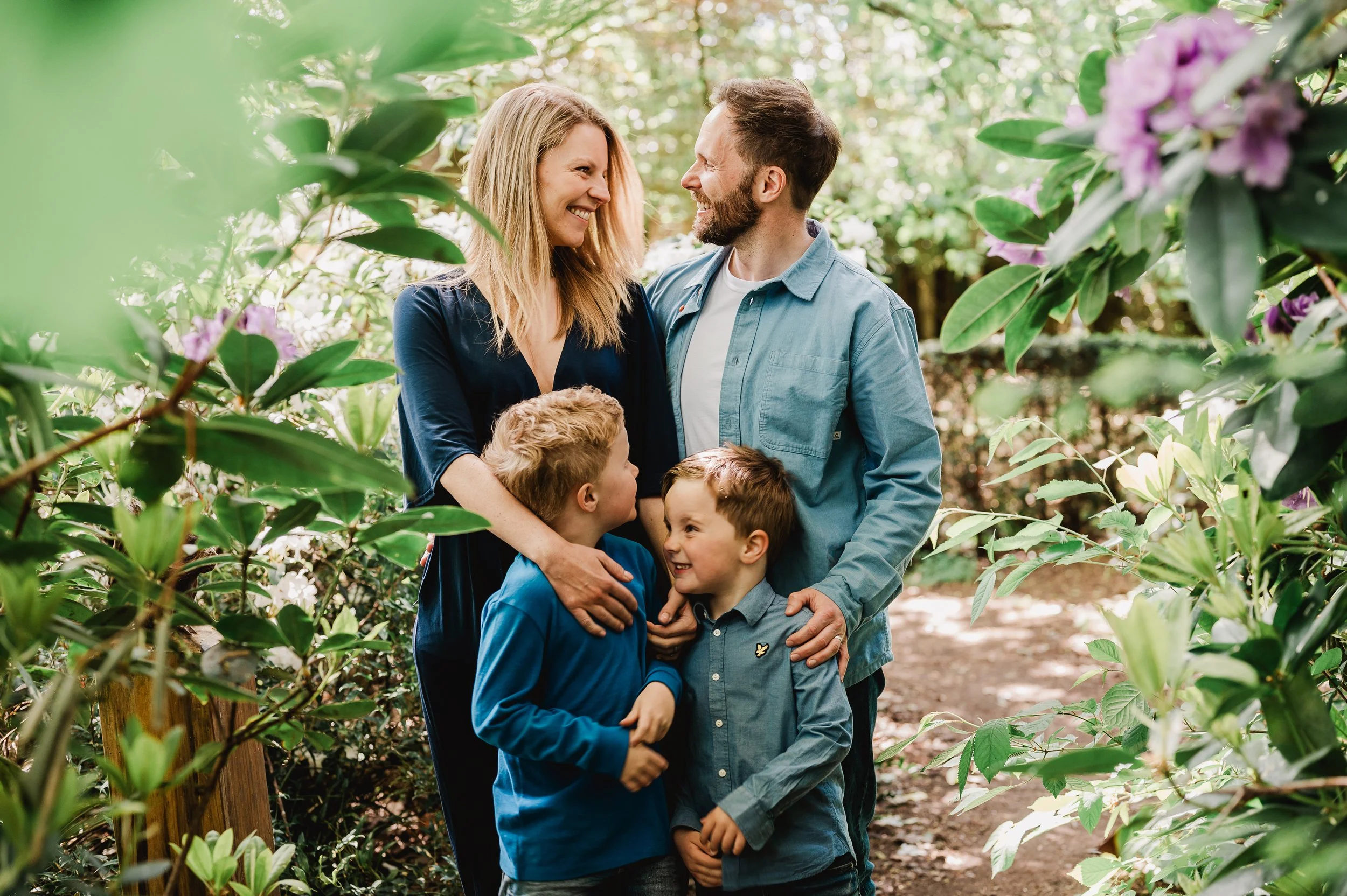 A happy family of four, including a mother, father, and two young boys, smiling and embracing each other in a lush, green garden with flowering bushes and sunlight filtering through the trees.