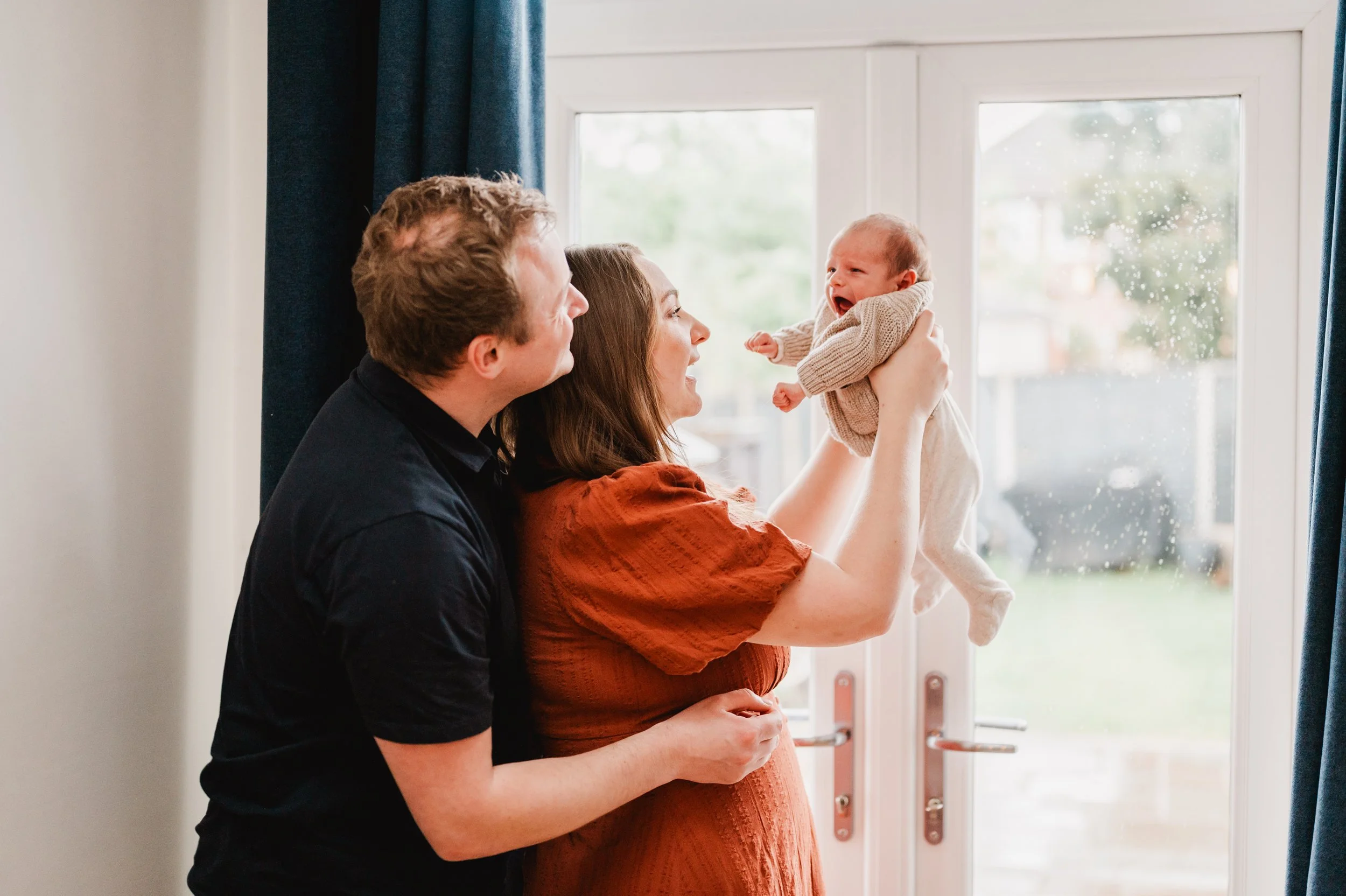 A family with a man, woman, and baby standing by a window; the woman is holding the baby, who is crying, and the man is smiling at them.