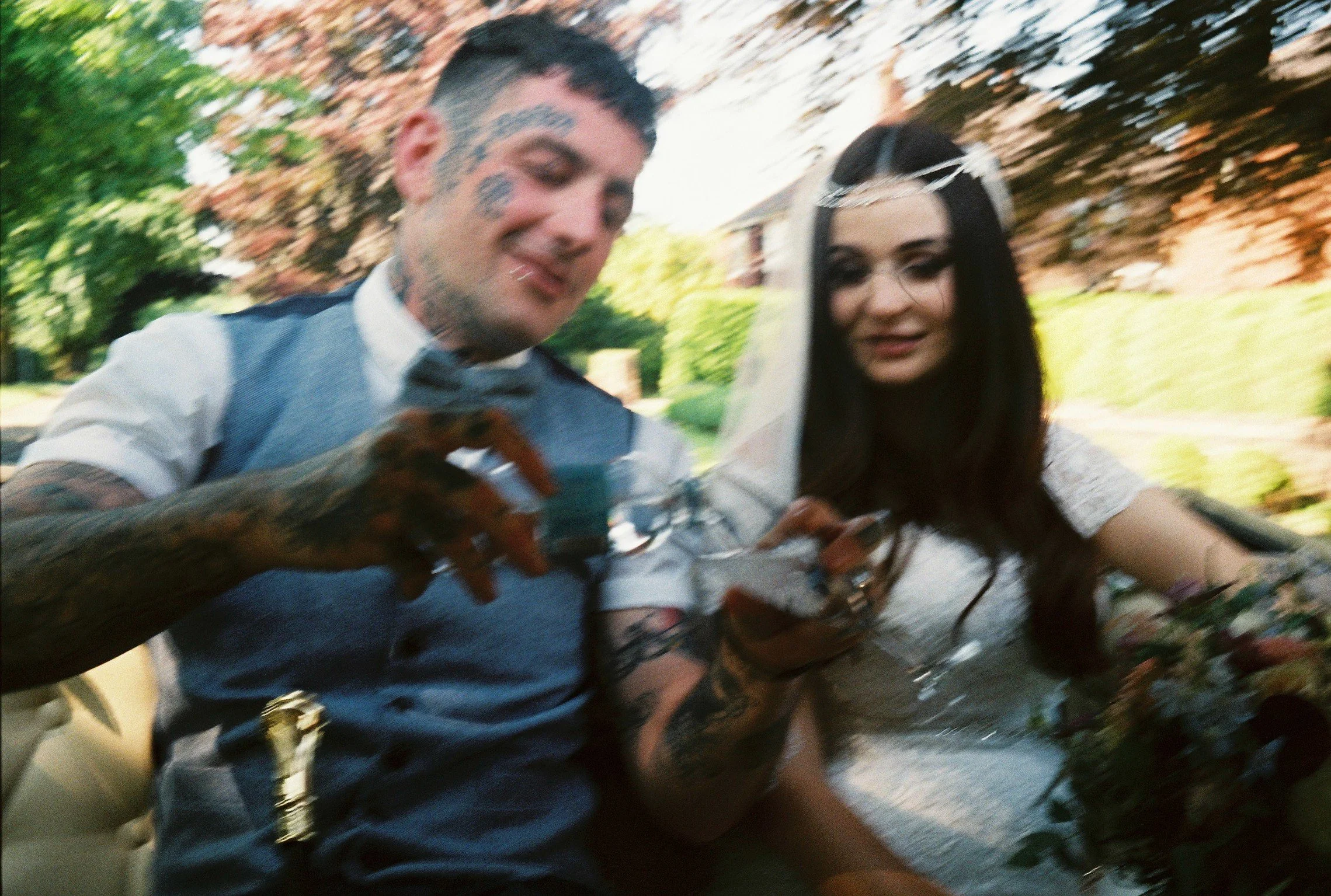 A tattooed man and a woman in a wedding dress look at their phones outdoors with greenery and trees in the background.
