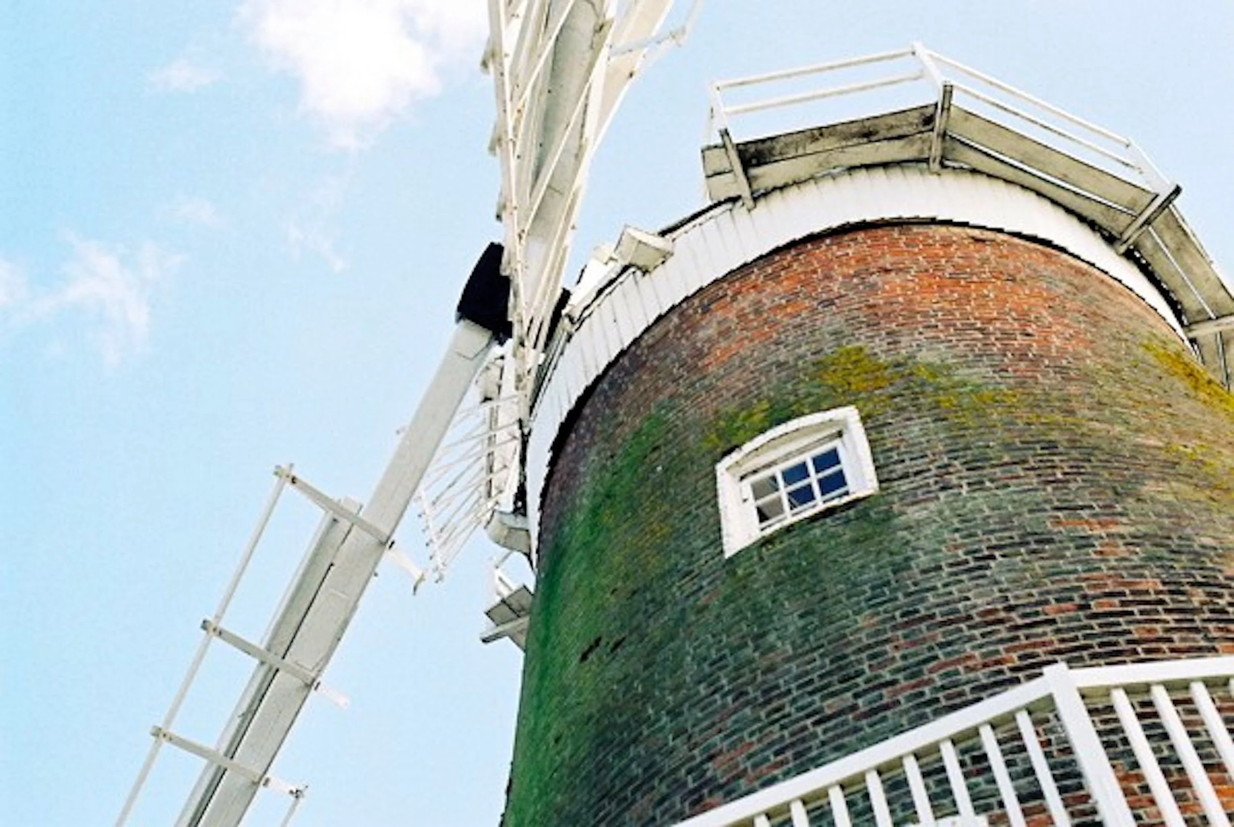 A low-angle view of a brick windmill tower with a white balcony and a white wooden ladder, against a blue sky with some scattered clouds.