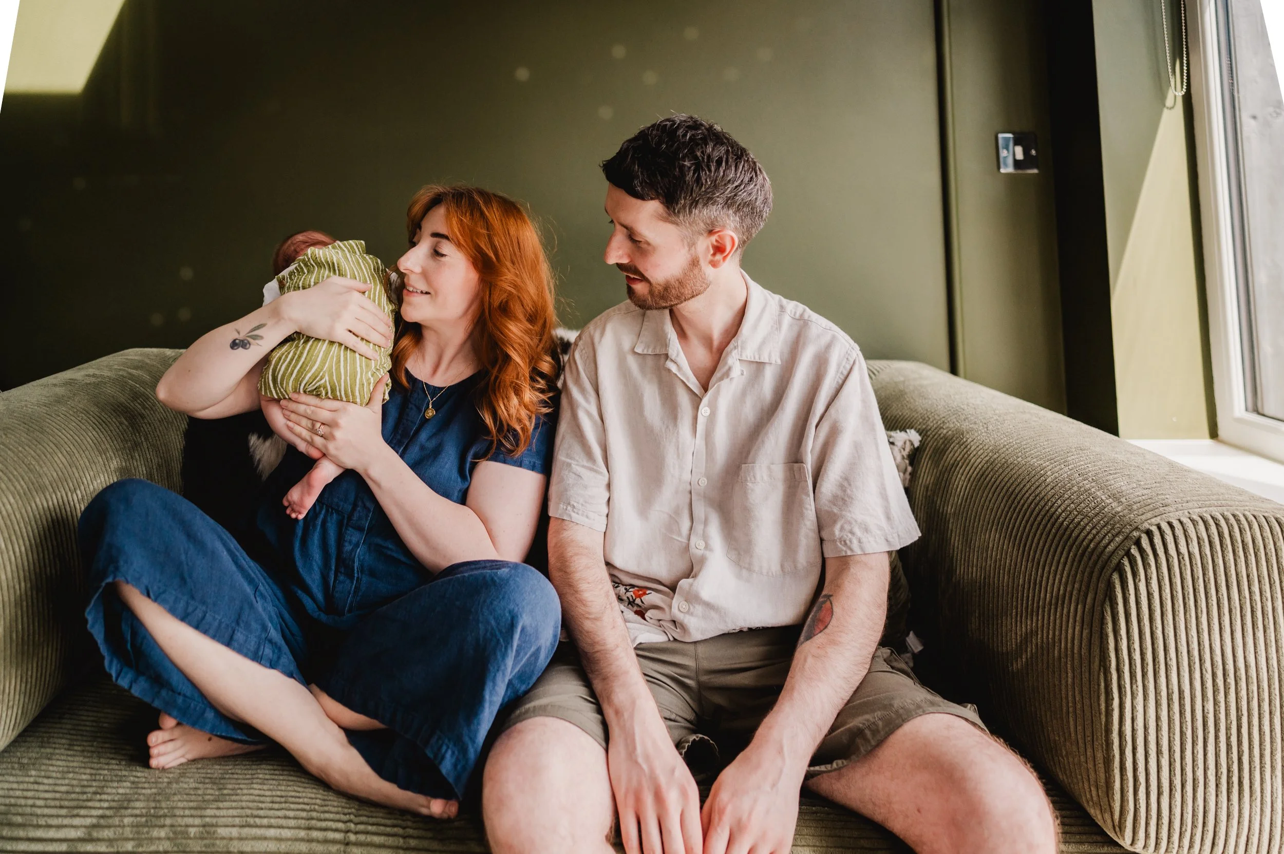 A woman with red hair holding a baby while sitting on a couch, accompanied by a man with short brown hair. They are in a cozy room with a green wall and a window on the right. The woman looks fondly at the baby, and the man looks at the woman.