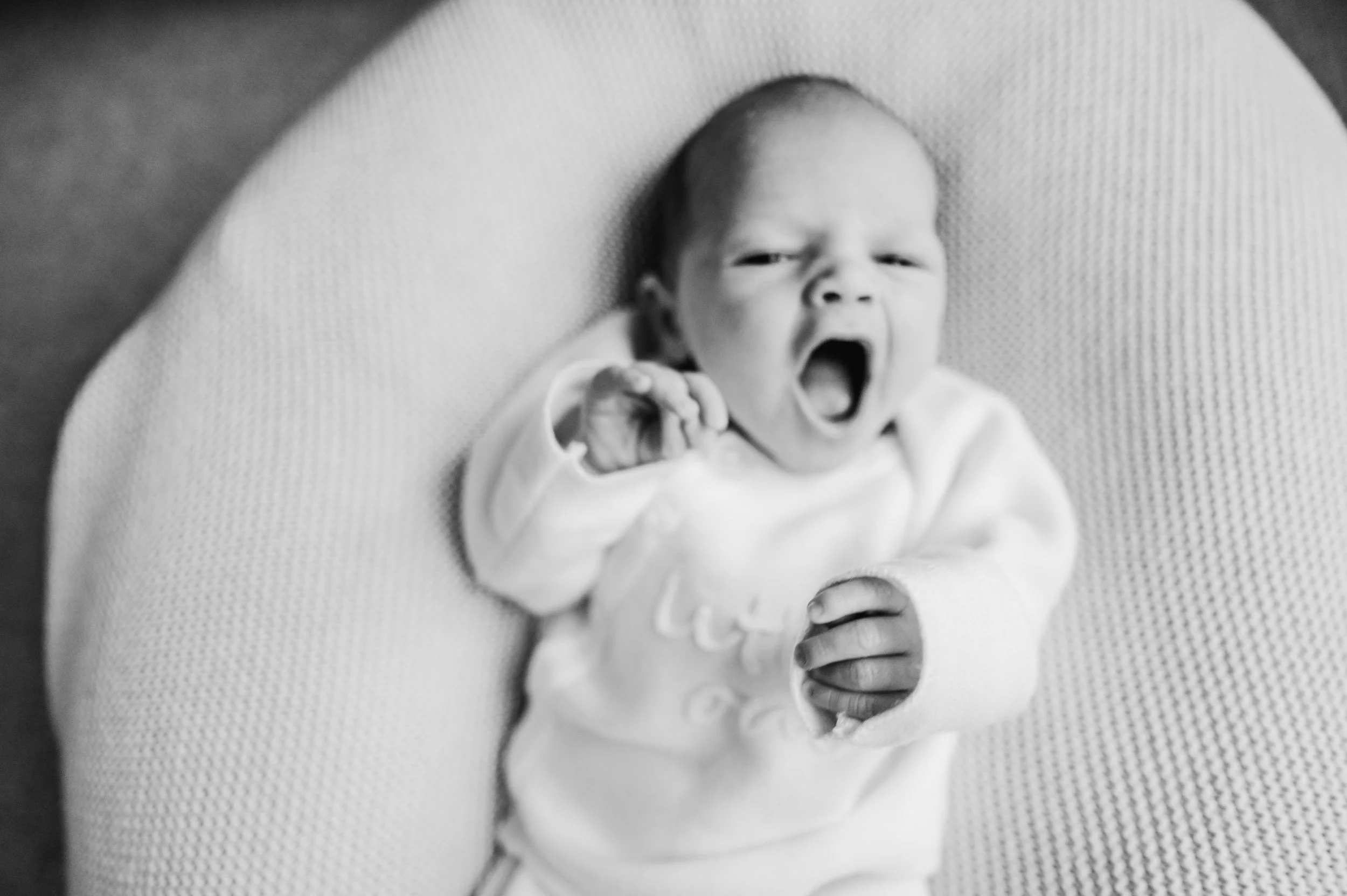 Black and white photo of a yawning baby lying on a padded surface, wearing a white outfit, with one hand near the face.