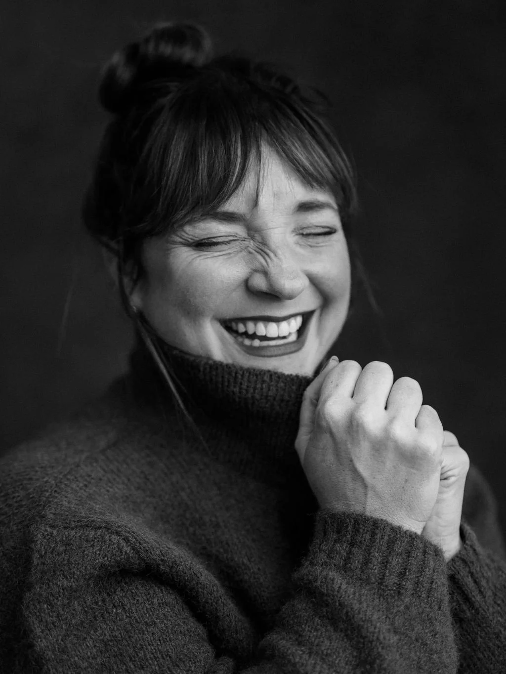 Black and white close-up of a woman laughing, eyes closed, with her hands clasped near her chin, wearing a turtleneck sweater and having her hair styled in an updo.