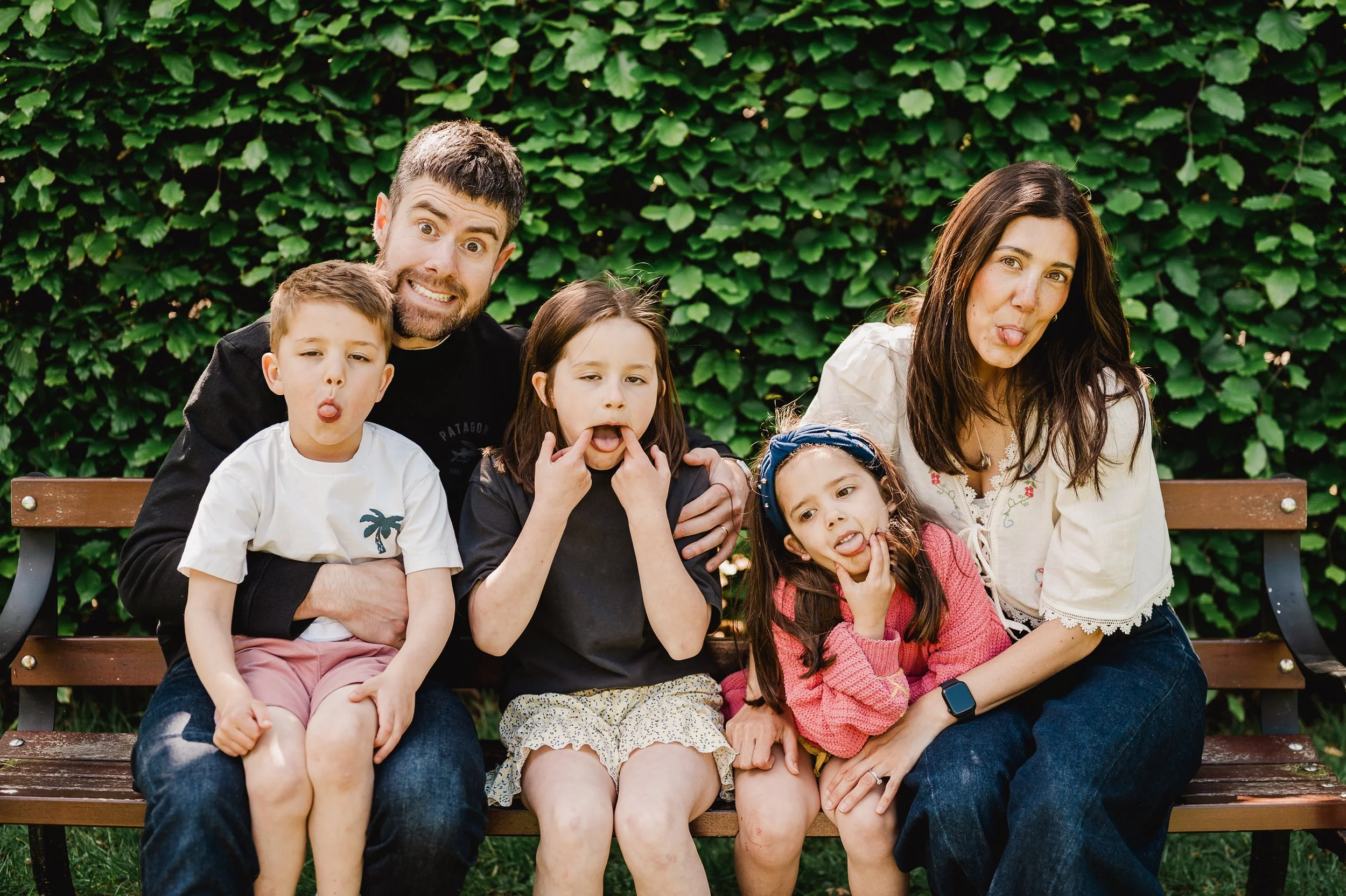 A family of six sitting on a wooden park bench in front of green leafy bushes. The adults appear to be a man and a woman, with four children. They are making funny faces, with some sticking out their tongues and others pulling their lips.