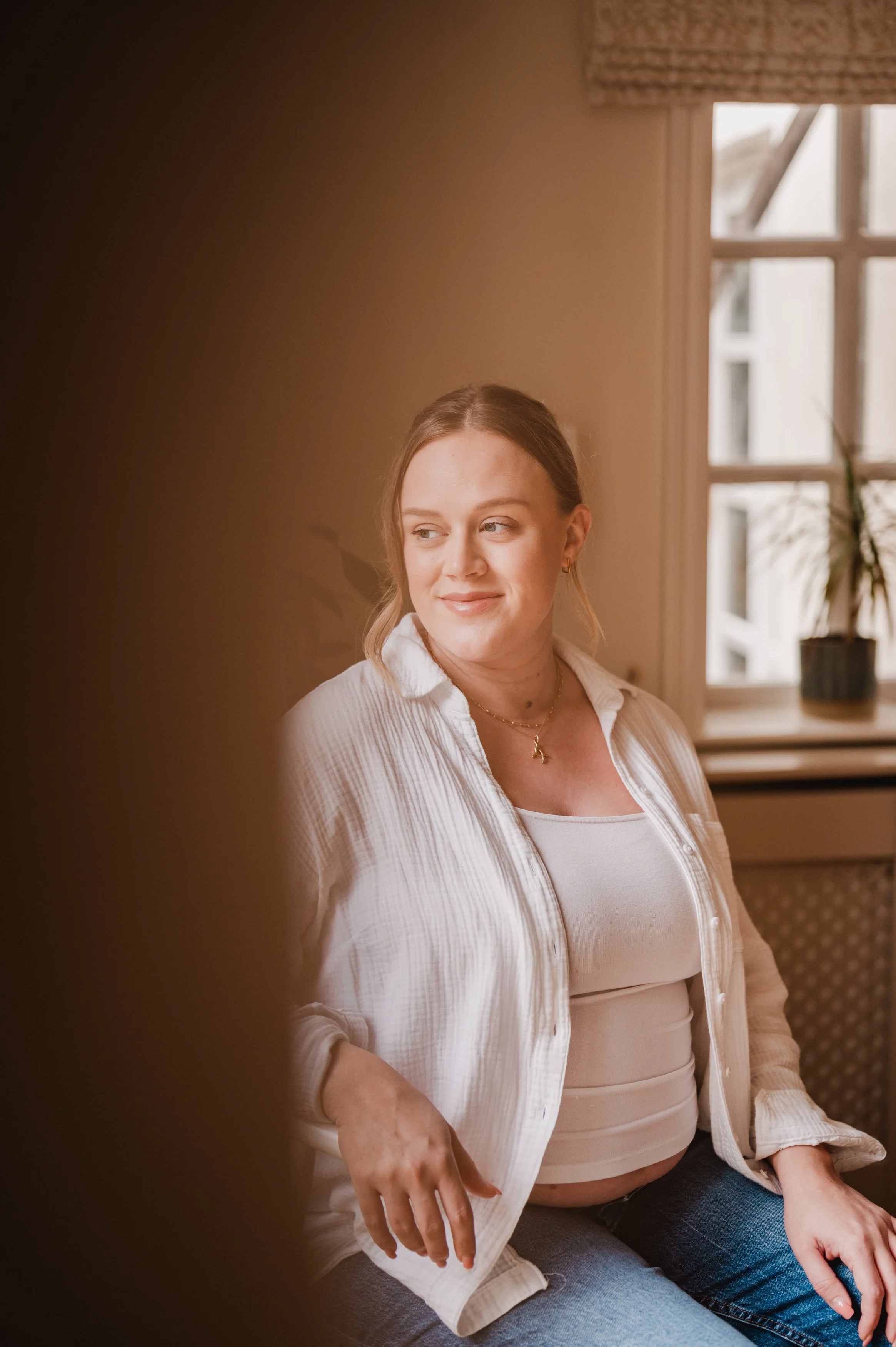 A woman sitting indoors by a window with natural light, wearing a white tank top and a light-colored, striped button-up shirt, with jewelry including earrings and a necklace.