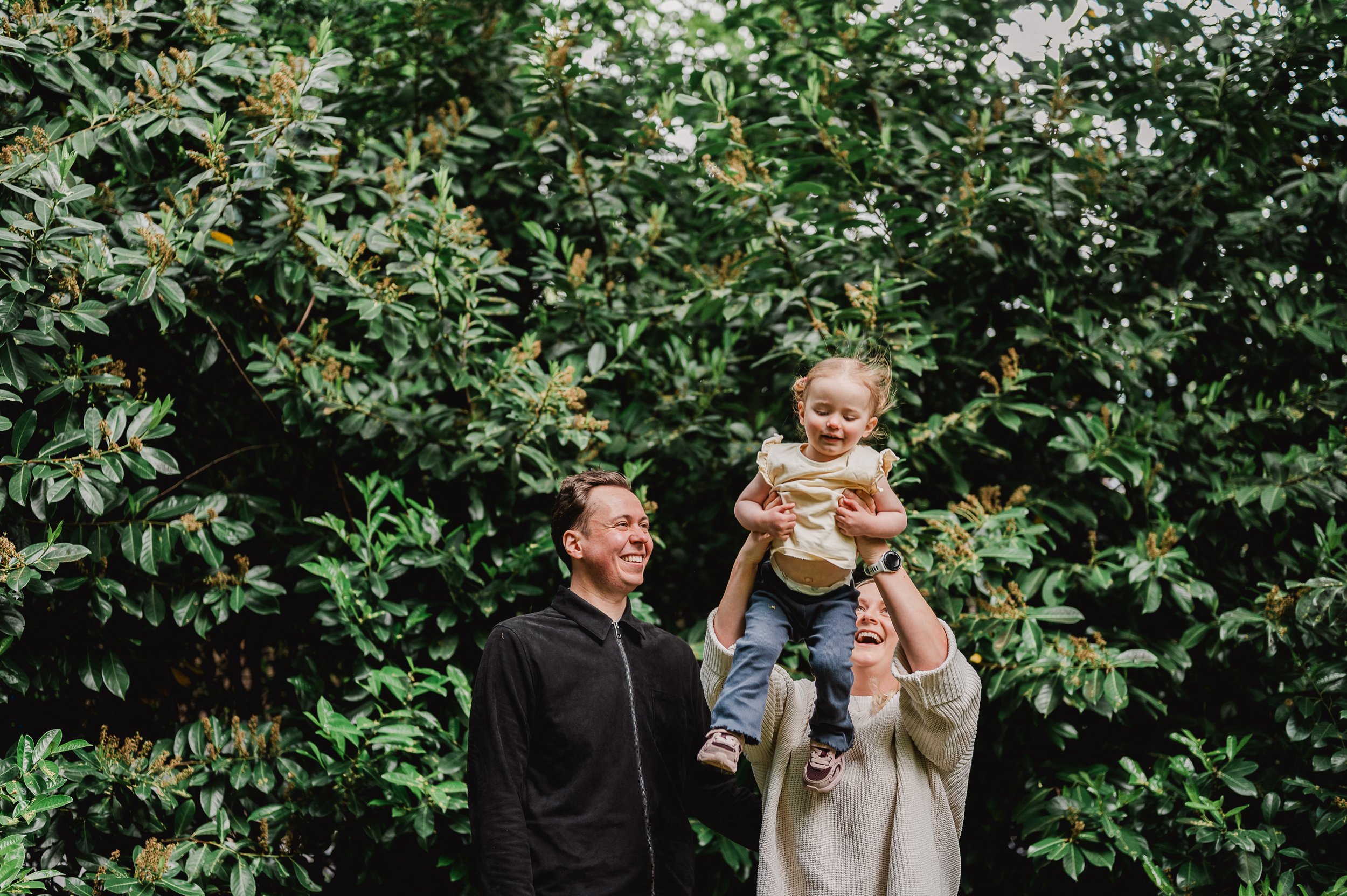 Happy family outdoors, young child being lifted by parents near lush green plants.
