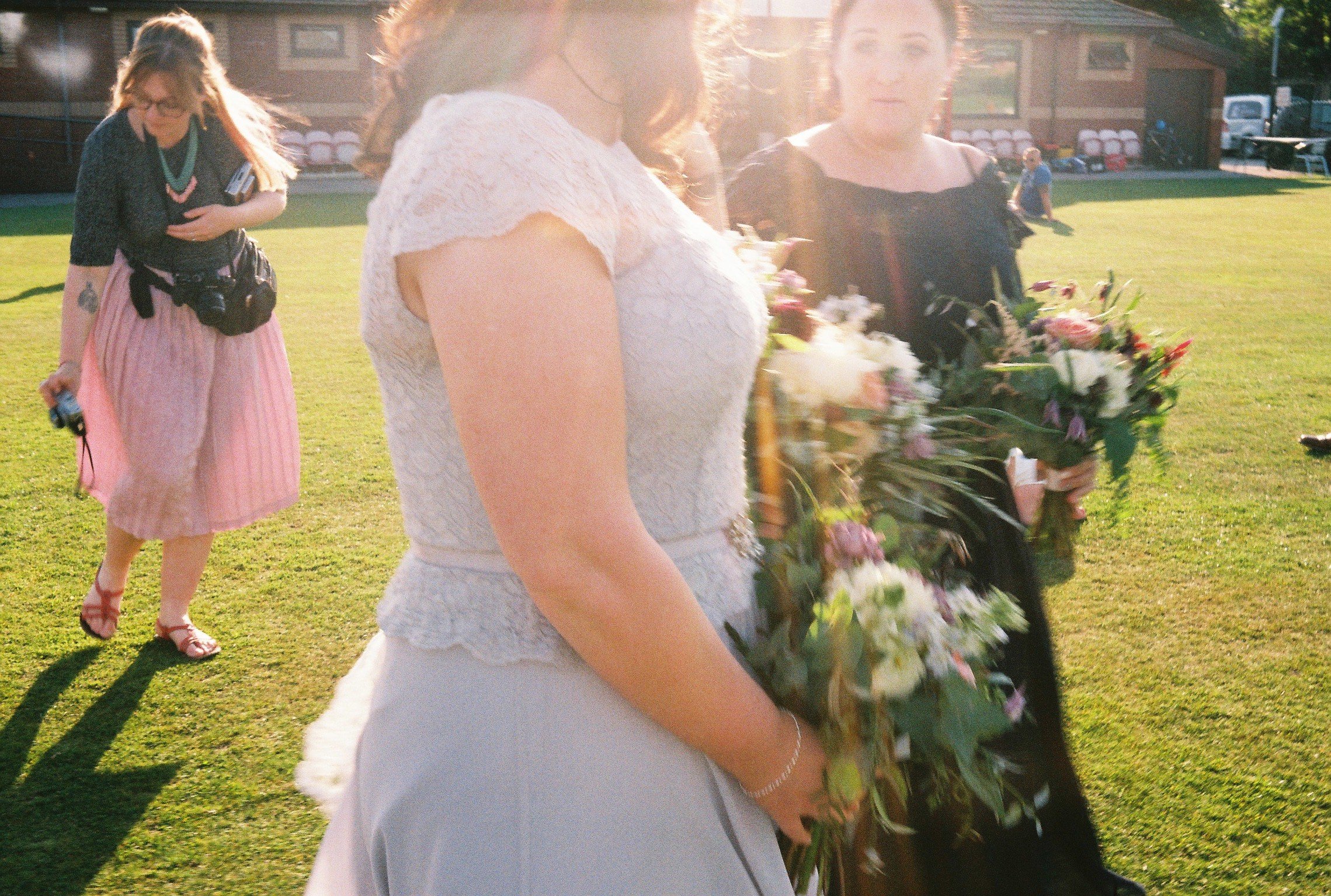 Women carrying bouquets of flowers on a grassy field during sunset, with a building and several chairs in the background.
