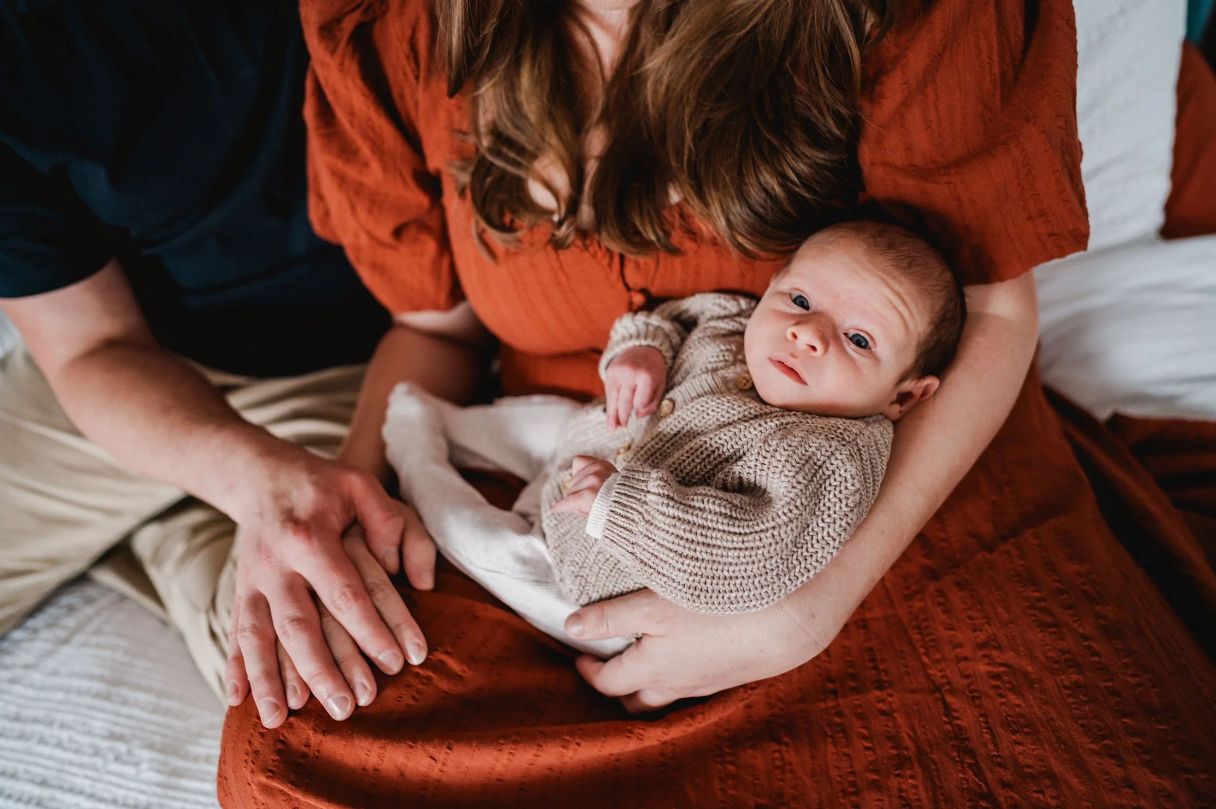 A person holding a baby inside, with only the person's arms and part of their torso visible. The person is wearing a rust-colored, textured shirt, and the baby is dressed in a beige, knit sweater with small buttons. The baby has wide eyes and is look