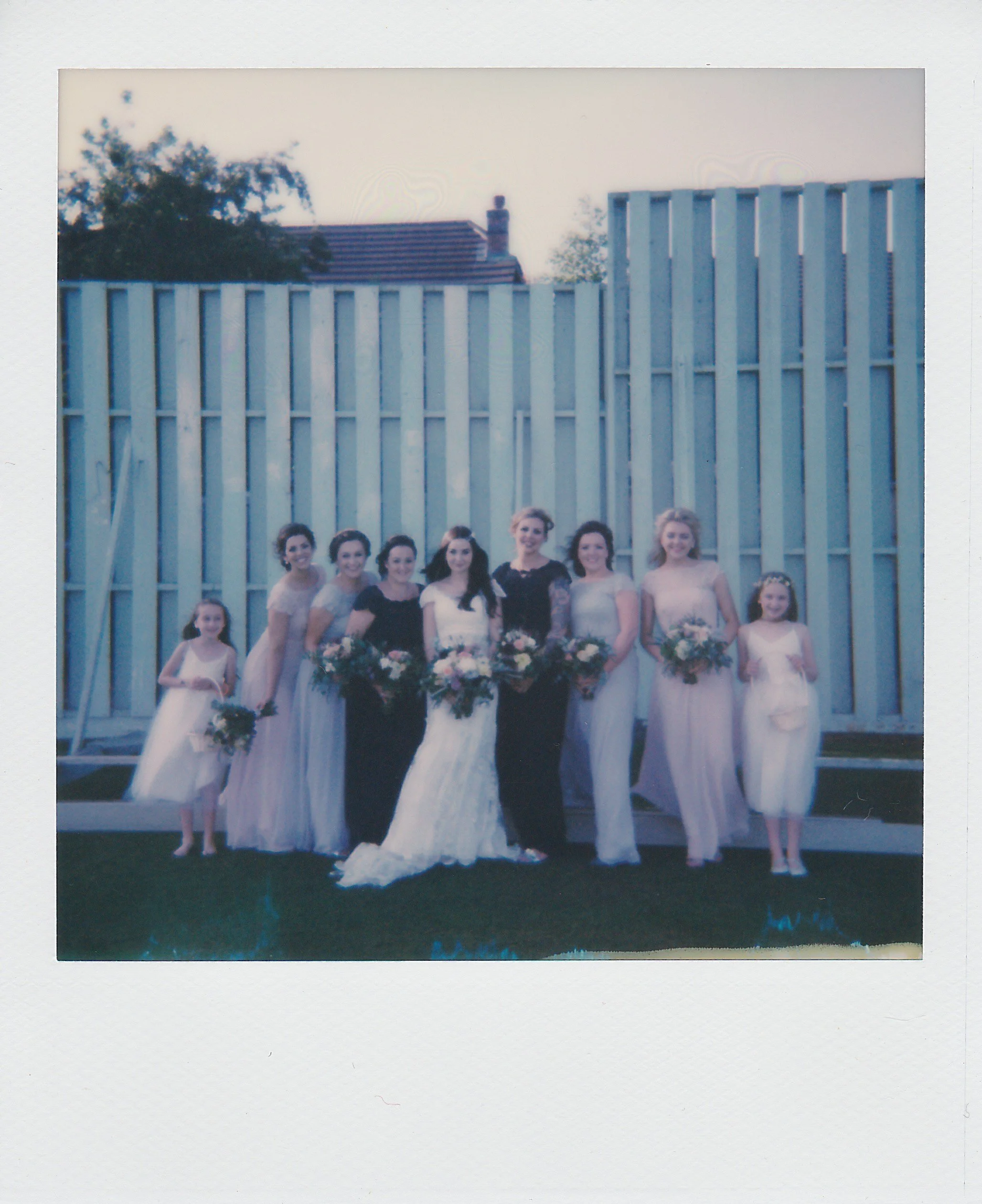 Group of women in formal dresses and young girls in white dresses, holding bouquets, posing outside in front of a blue fence, at a wedding celebration.