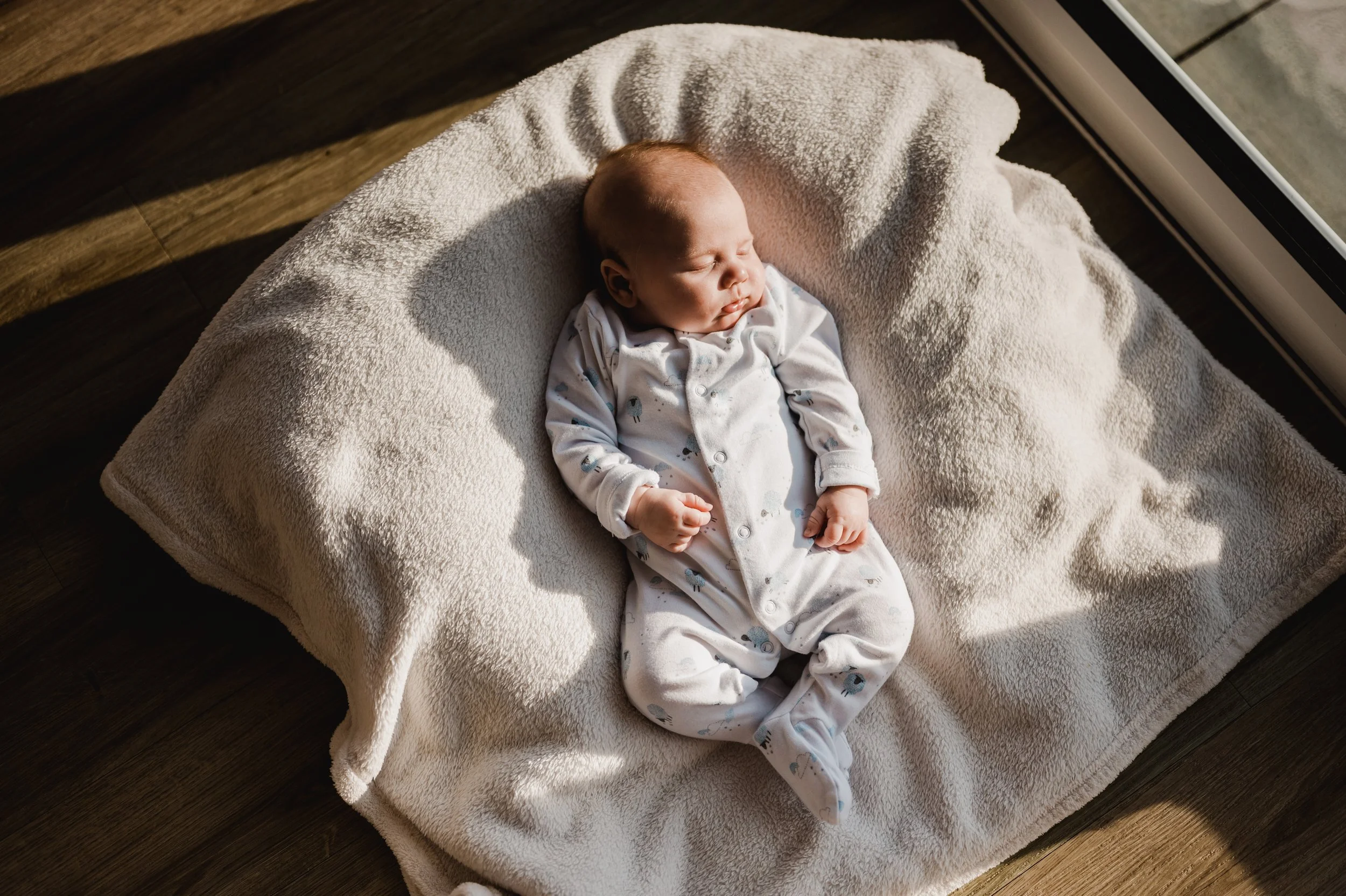 A sleeping baby lying on a soft, fluffy blanket on a wooden floor near a window.