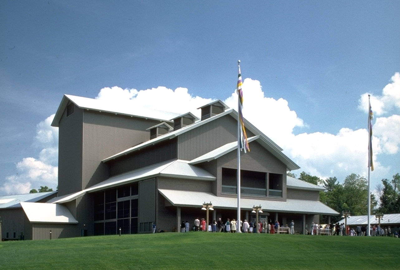 PROJECT: Alice Busch Opera Theater LOCATION: Cooperstown, New York CLIENT: Glimmerglass Opera Company COMPLETION: 1987 SIZE: 12,000 Square Feet COST: $4 Million The Alice Busch Opera Theater, designed in 1987 by Hugh Hardy for the renowned Glimmerglass Opera Company, presents sophisticated performances in an informal, comfortable, intimate setting. The theater was carefully sited to take full advantage of its 20-acre arcadian setting. Patrons arrive on terraced parking levels and reach the theater by walking across a meadow and around a pond. The curving path leads past an open courtyard to the outdoor entrance lobby. From here, the parking area disappears into the landscape, preserving the tranquility of the scene. The exterior has little precedent as a structure for music, because its character is derived from surrounding farm buildings, several of which are now used for theater support functions. These structures are recalled in its sloping roofs and massing and in its profiled wood and metal textures. The opera house is an open-air pavilion with screened side walls and rolling doors that can be opened or closed for matinees, inclement weather, or natural ventilation, revealing views of the surrounding landscape. All of the hall’s 900 seats are within 70 feet of the stage. The large stage and high fly loft provide ample space for handling scenery. The orchestra pit accommodates 80 musicians.