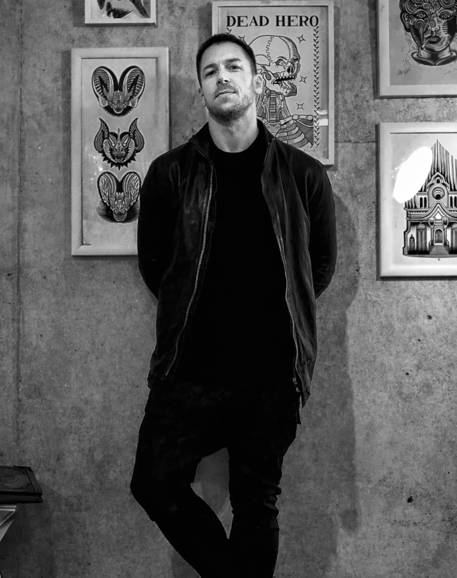 A man standing in front of a wall with various wall art prints, including a skull labeled "Dead Hero" and multiple animal-style portraits. He is wearing a black jacket and black pants, with his hands in his pockets, looking at the camera.