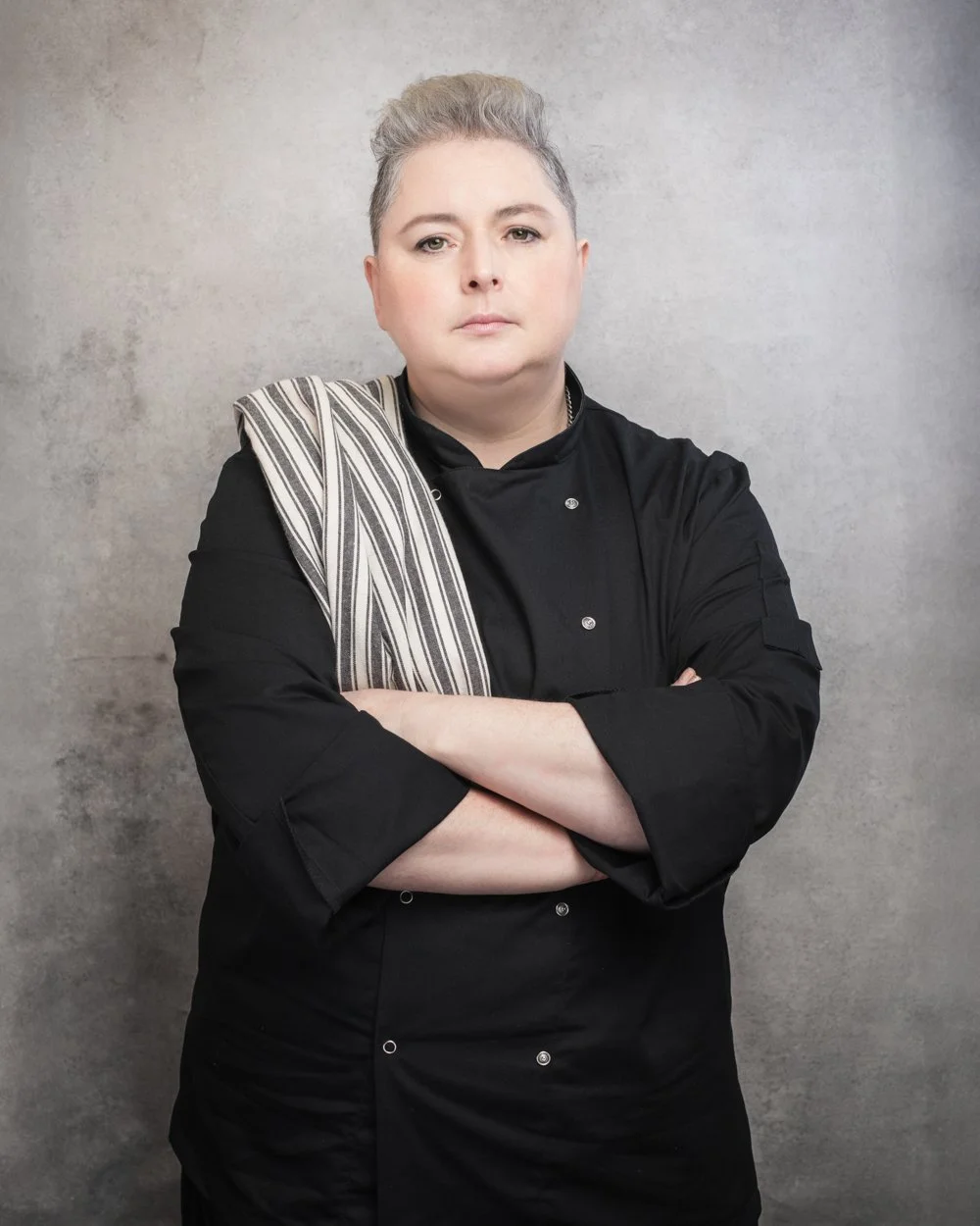 A confident female chef with short, light gray hair, wearing a black chef's coat and a striped apron over her shoulder, standing against a gray textured background with her arms crossed.