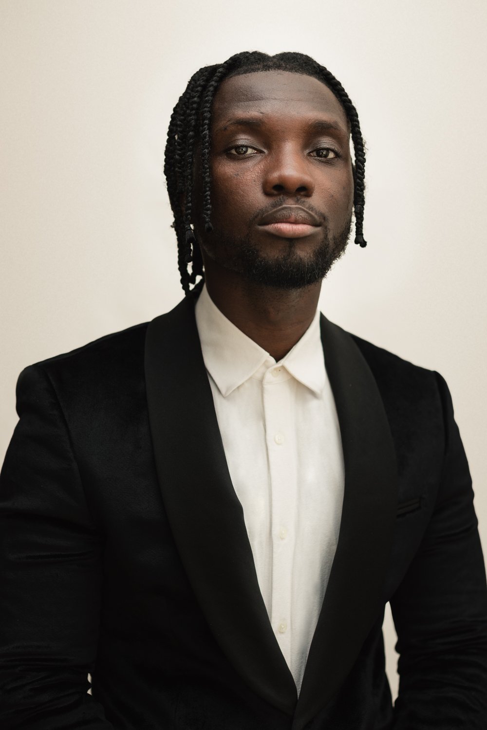 A young man with dark skin and styled braids, wearing a white dress shirt and a black blazer, looking at the camera with a neutral expression against a plain light background.