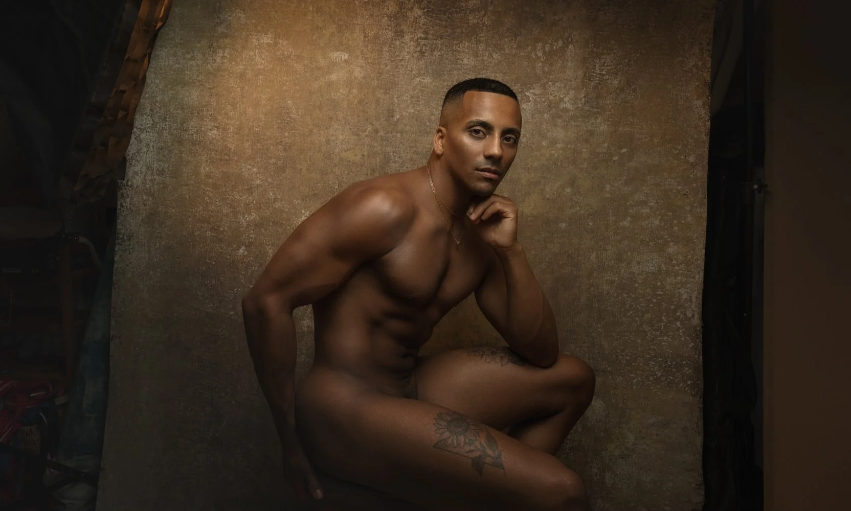Nude man with tattoos posing against a textured brown background, with one hand resting on his chin and looking at the camera.