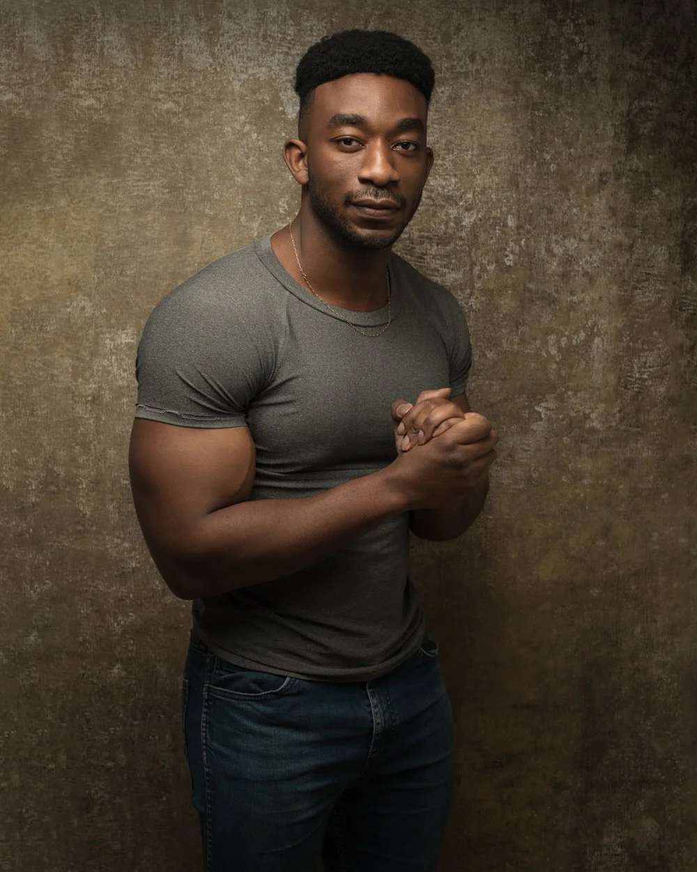 A young man with dark hair and a goatee, wearing a gray t-shirt and dark jeans, stands against a textured brown background, looking directly at the camera with his hands clasped in front of him.