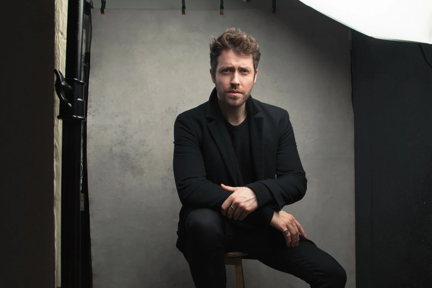 A man with brown hair and a beard, wearing a black blazer and black shirt, sitting on a stool against a plain gray background in a photography studio with lighting equipment visible.