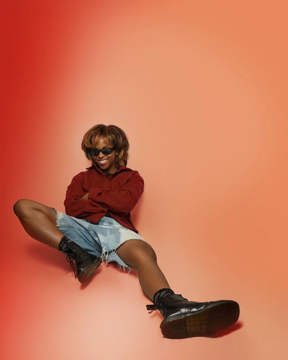 Young woman sitting on the floor with legs spread, wearing sunglasses, a red jacket, denim shorts, black combat boots, and smiling against a peach-colored background.