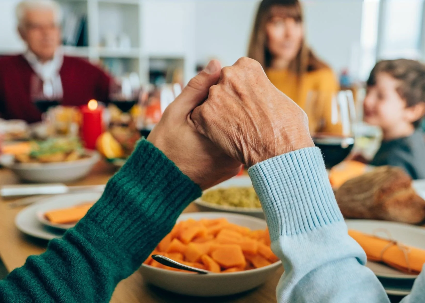 A family of all ages gathered around a table full of food. In the foreground are two holding hands.