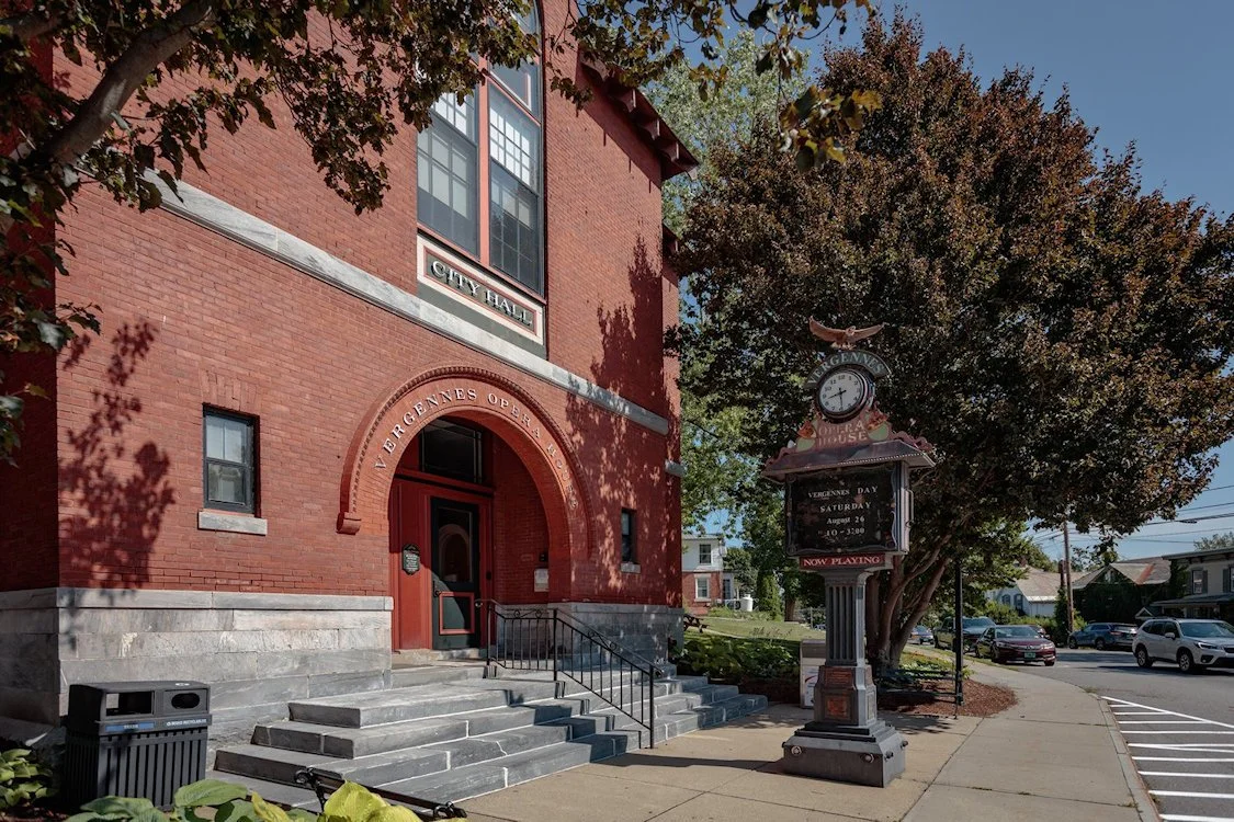 A red brick city hall building with a sign for Vergennes Opera House and a vintage clock on a post nearby, surrounded by trees and parked cars on the street.