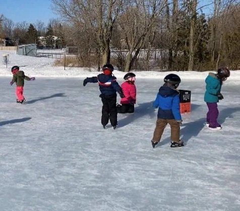 Children playing on an ice rink during winter with some trees and a fence in the background.