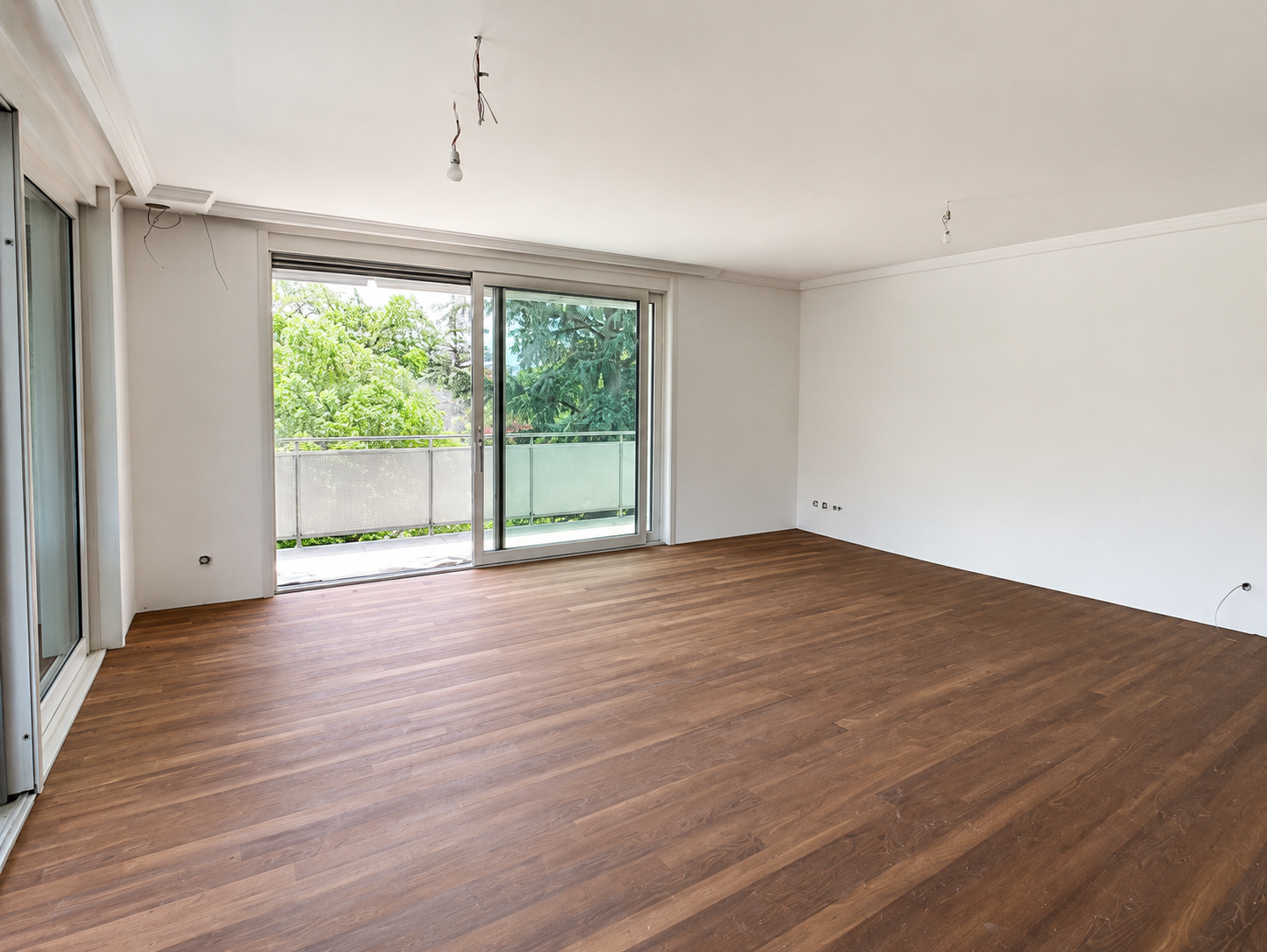 Empty living room with large sliding glass doors leading to a balcony, white walls, and wooden flooring.