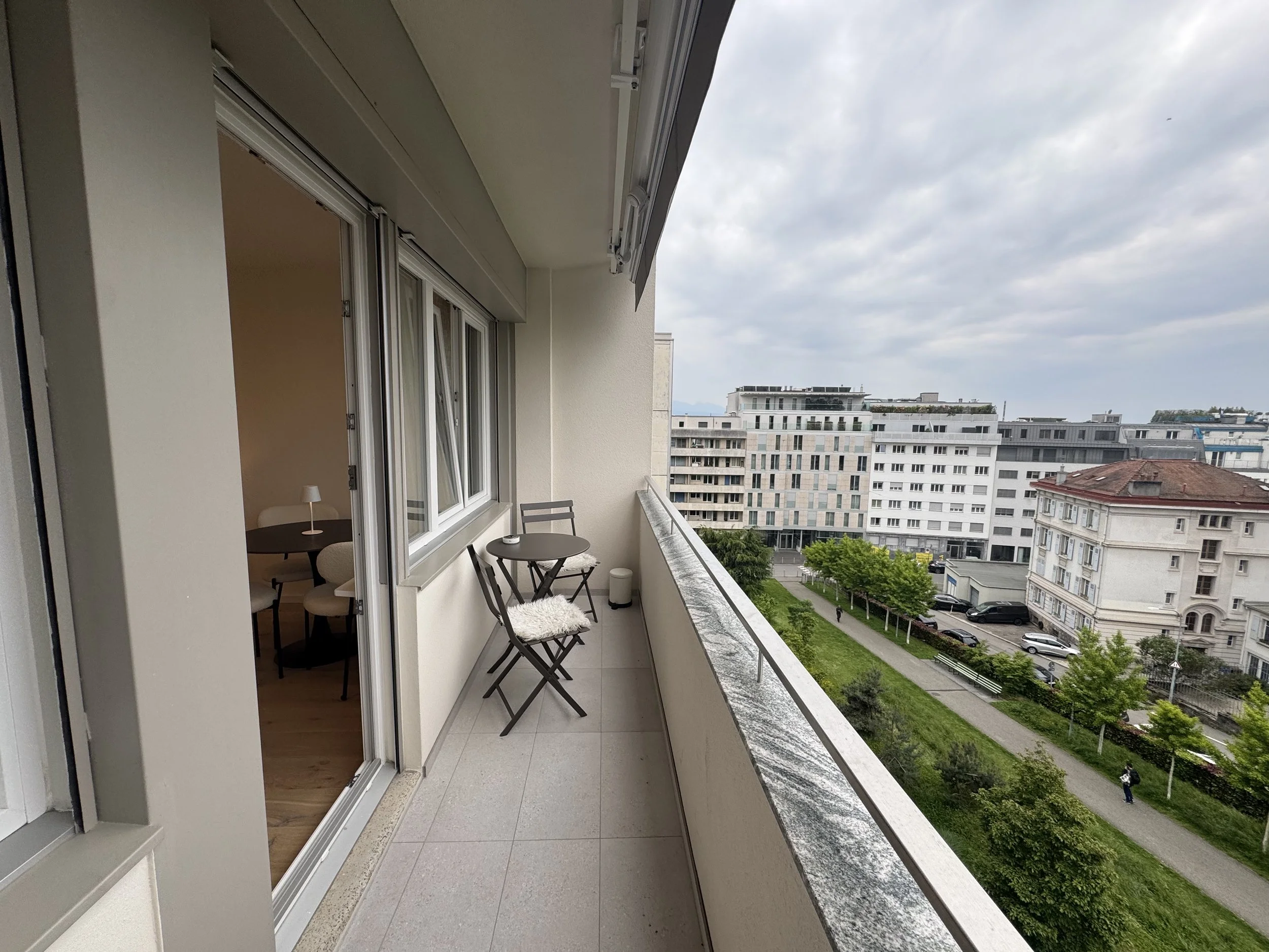 A balcony with a small round table and chairs, with a view of multiple modern apartment buildings and green trees under a cloudy sky.