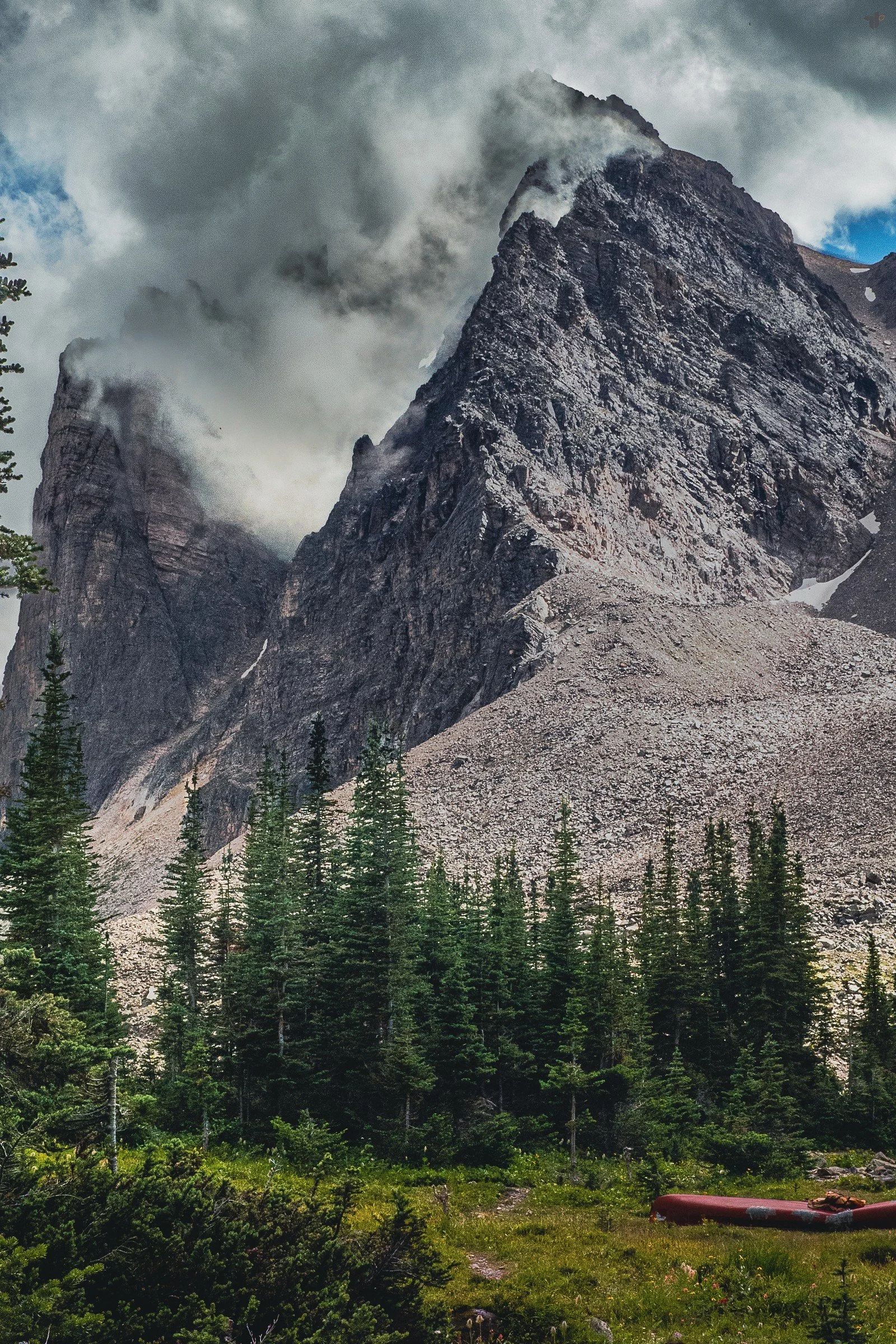 A steep rocky mountain rises into cloud above dark evergreen trees.