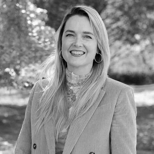 A smiling woman with long blonde hair, wearing a blazer and hoop earrings, standing outdoors with trees in the background.