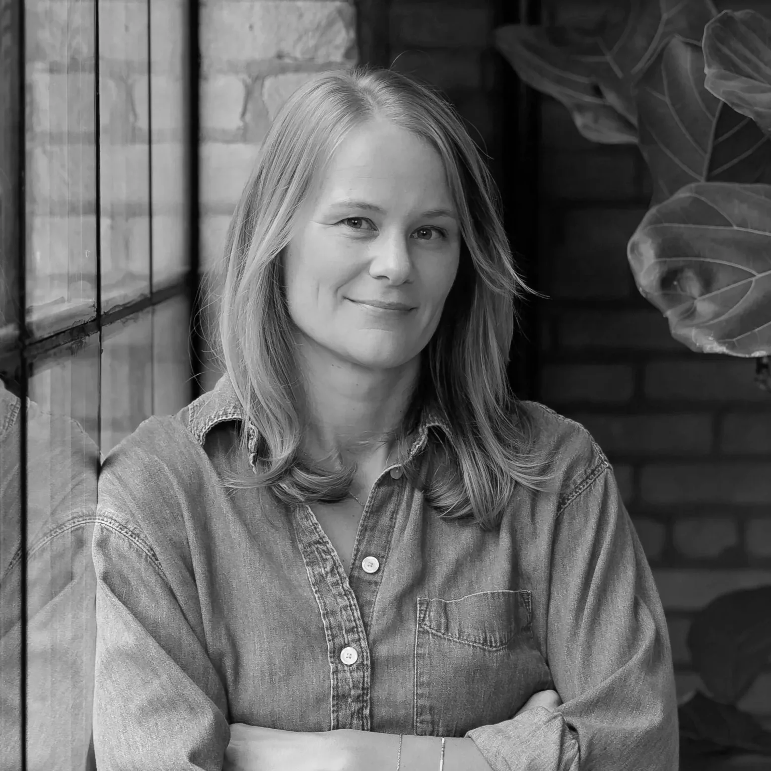 A black and white portrait of a woman with shoulder-length hair, wearing a denim shirt, smiling slightly, sitting indoors next to a window and large indoor plants.