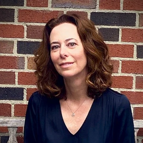 A woman with shoulder-length curly brown hair wearing a navy blue top standing in front of a red brick wall.