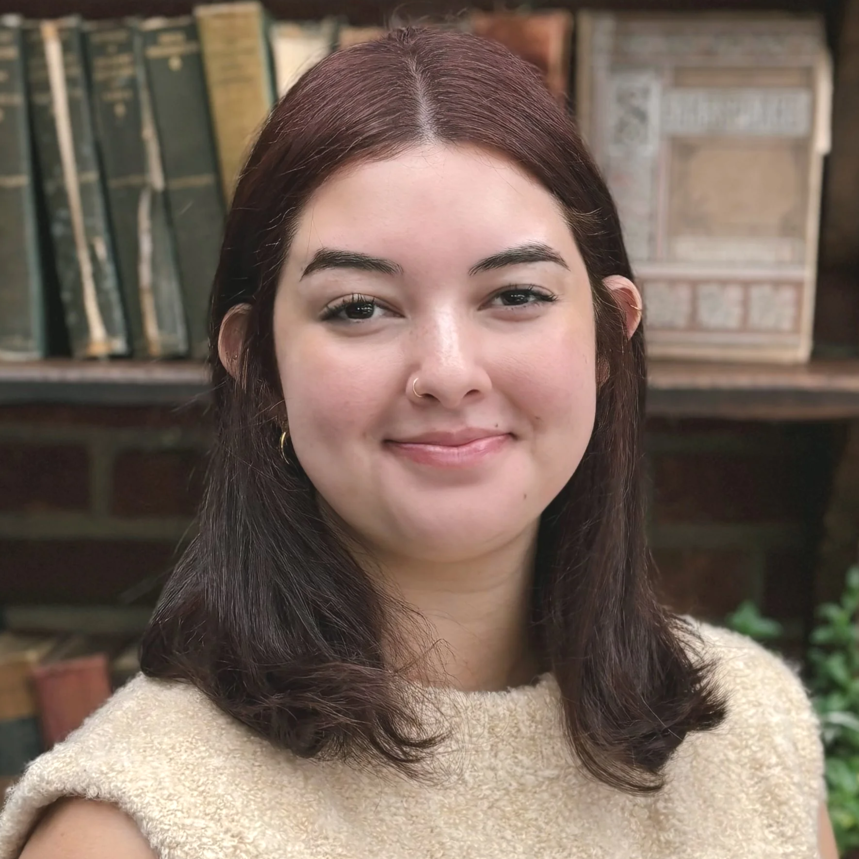 A woman gottman couples therapist with shoulder-length brown hair, wearing a beige sleeveless top, smiling in front of a bookshelf filled with old books.