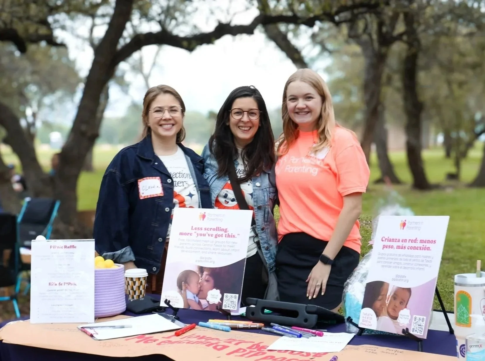 Three women smiling at an outdoor event, standing behind a table with informational signs, pens, and raffle prizes, with trees in the background.