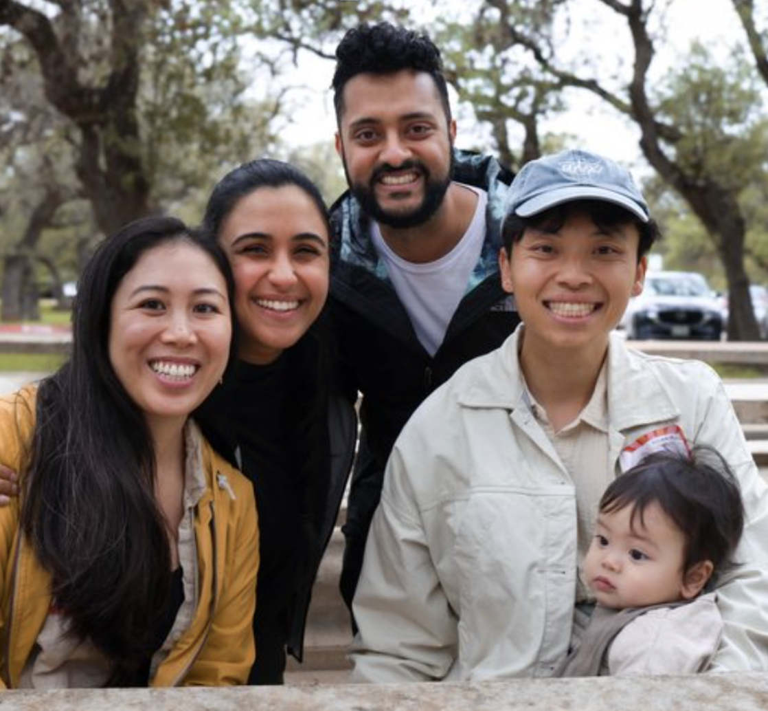 Group of five people smiling outdoors in a park, with trees and cars in the background, three women and two men, one of the women is holding a young child.