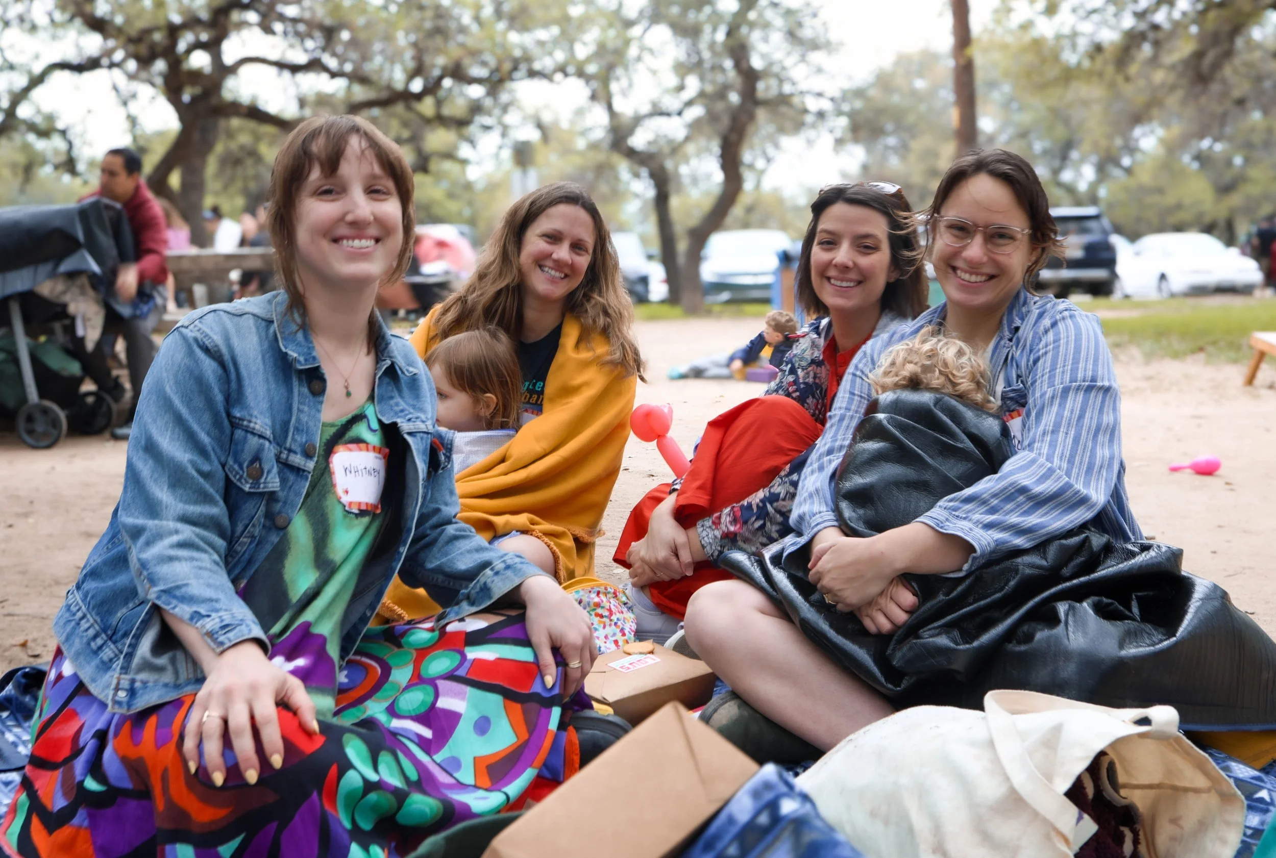 Four women sitting outdoors on the ground, smiling. Behind them are trees and other people, some standing and some sitting, with cars parked in the background. It appears to be a park or outdoor gathering.