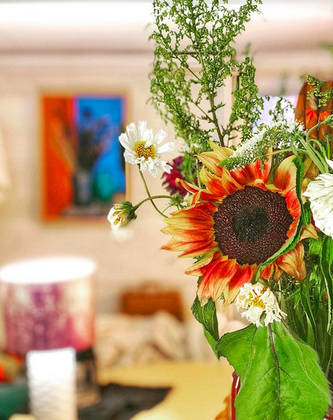 A photograph of a bedroom focused on a bouquet of flowers slightly past their best. The rest of the room is in soft focus, but you can make out a lamp on a dresser, and a painting of a vase of flowers on the wall.