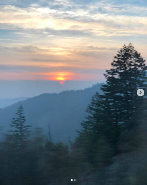 A hazy landscape photograph of mountains covered in forests, with a bright orange spot in the distance almost like a sunset.