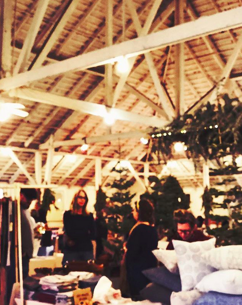 A soft focus photo of people milling in a booth at a holiday shopping bazaar, taking place in an old barn with high ceilings and exposed beams. Two women chat, a couple more people browse. There are wreaths and evergreen trees around.