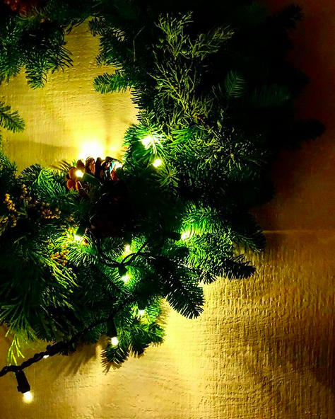 Green evergreen Christmas wreath at night with twinkle lights lit and acorns in it, against a yellow wall.
