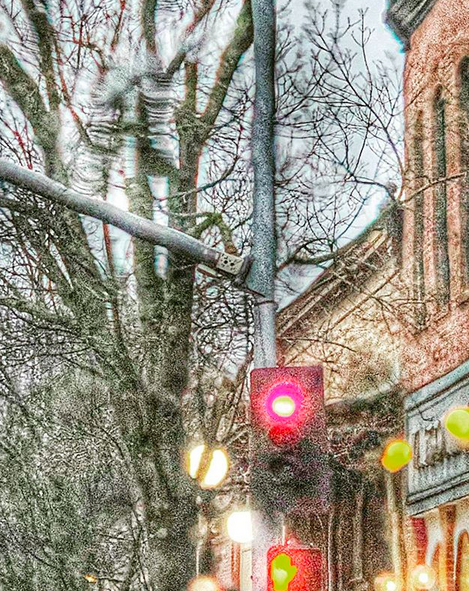 A rainy stop light scene, with bare trees, some shop fronts, and streetlights. Taken driving back from a client’s house.