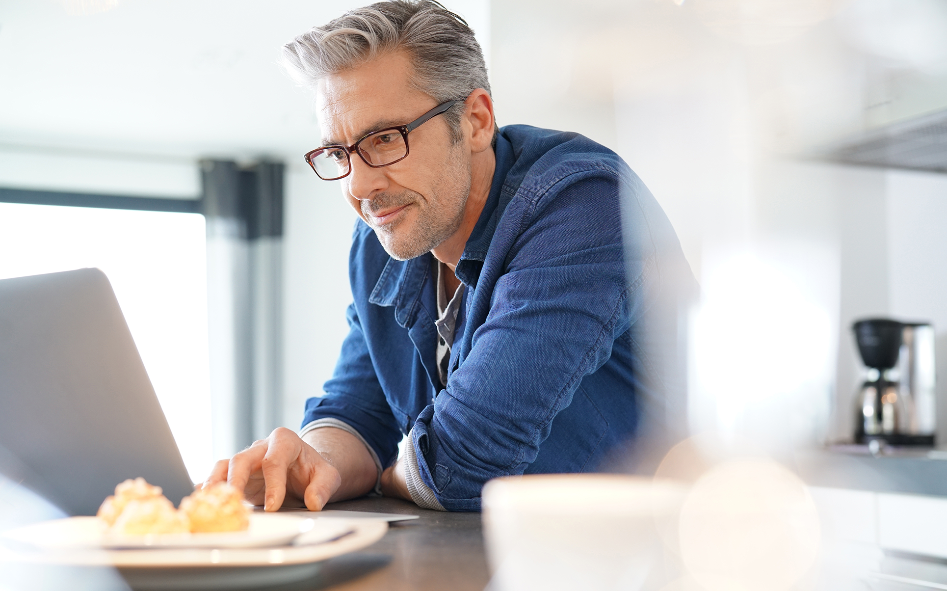 A middle-aged man with gray hair, glasses, and a beard, wearing a blue denim shirt, sits at a kitchen counter working on a laptop. There is a plate with some pastries or muffins in front of him. The setting appears to be a bright modern kitchen with a coffee maker in the background.