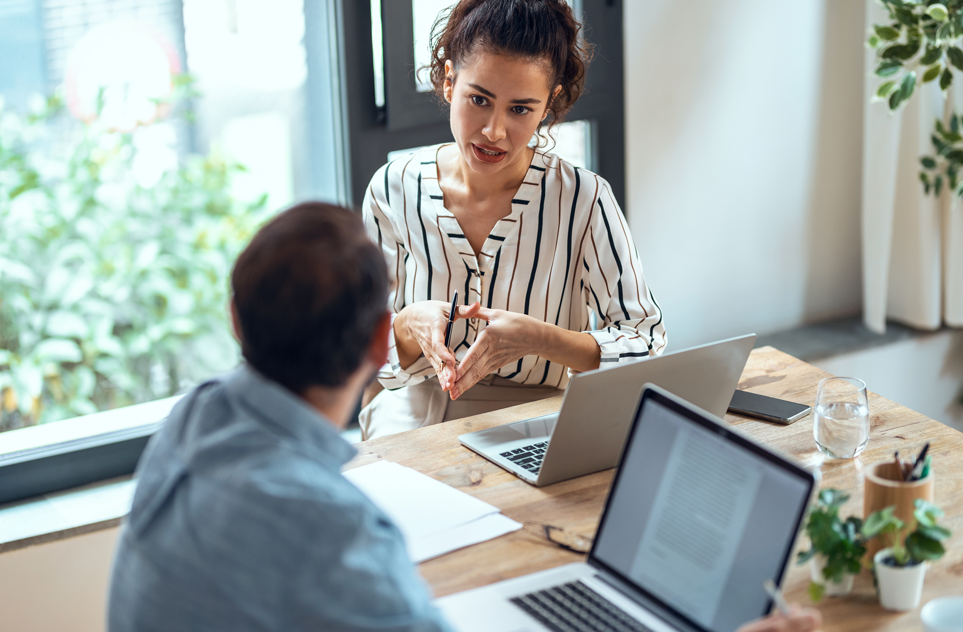 A woman with curly hair and a striped blouse is talking and explaining something to a man while sitting at a wooden table, which has laptops, a glass of water, a smartphone, a notebook, and a plant on it.